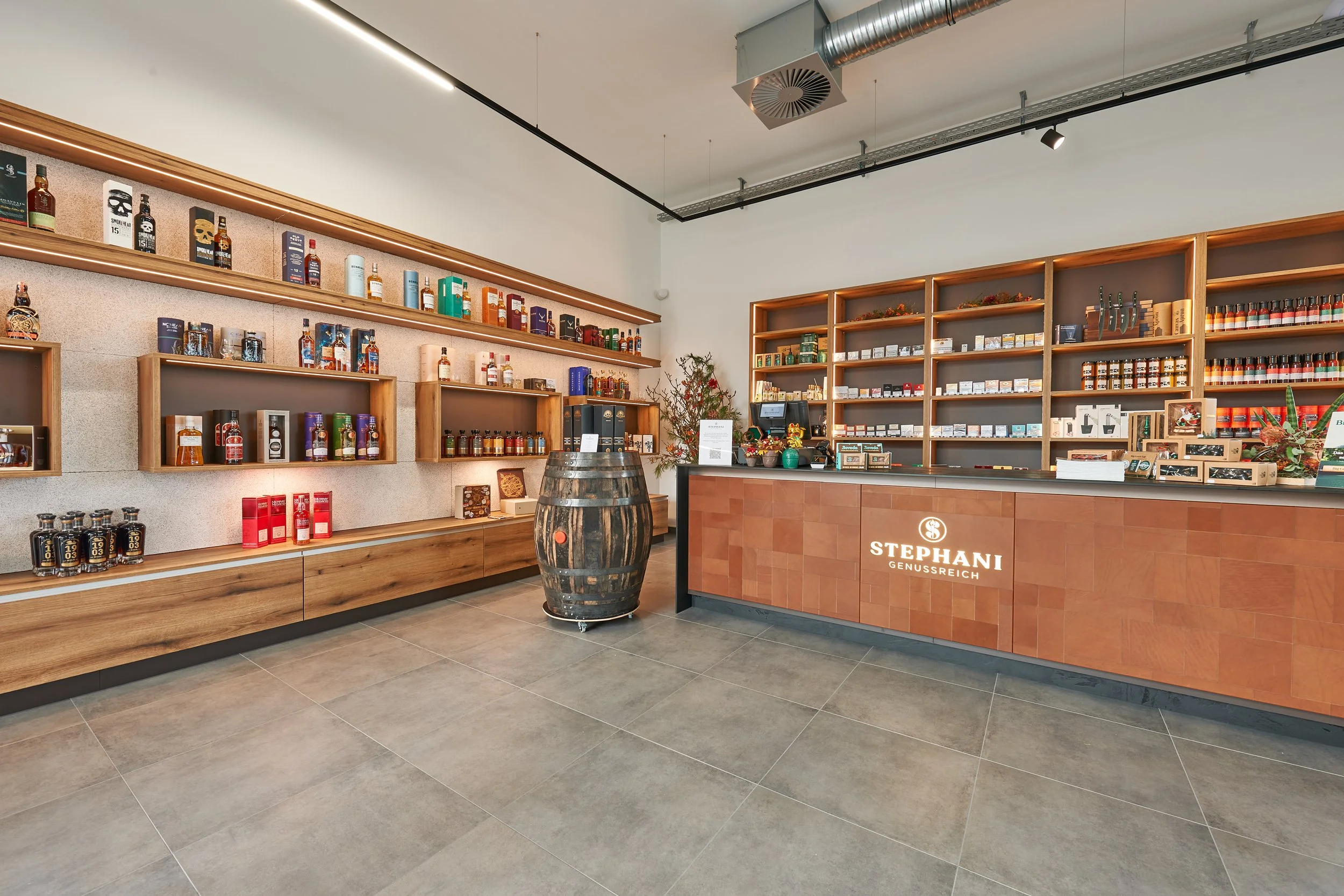 Innenansicht eines Geschenkeladens mit Holzregalen, die Spirituosen und andere Produkte präsentieren, und einem Tresen mit dem Schriftzug 'STEFANI GENUSSREICH'.