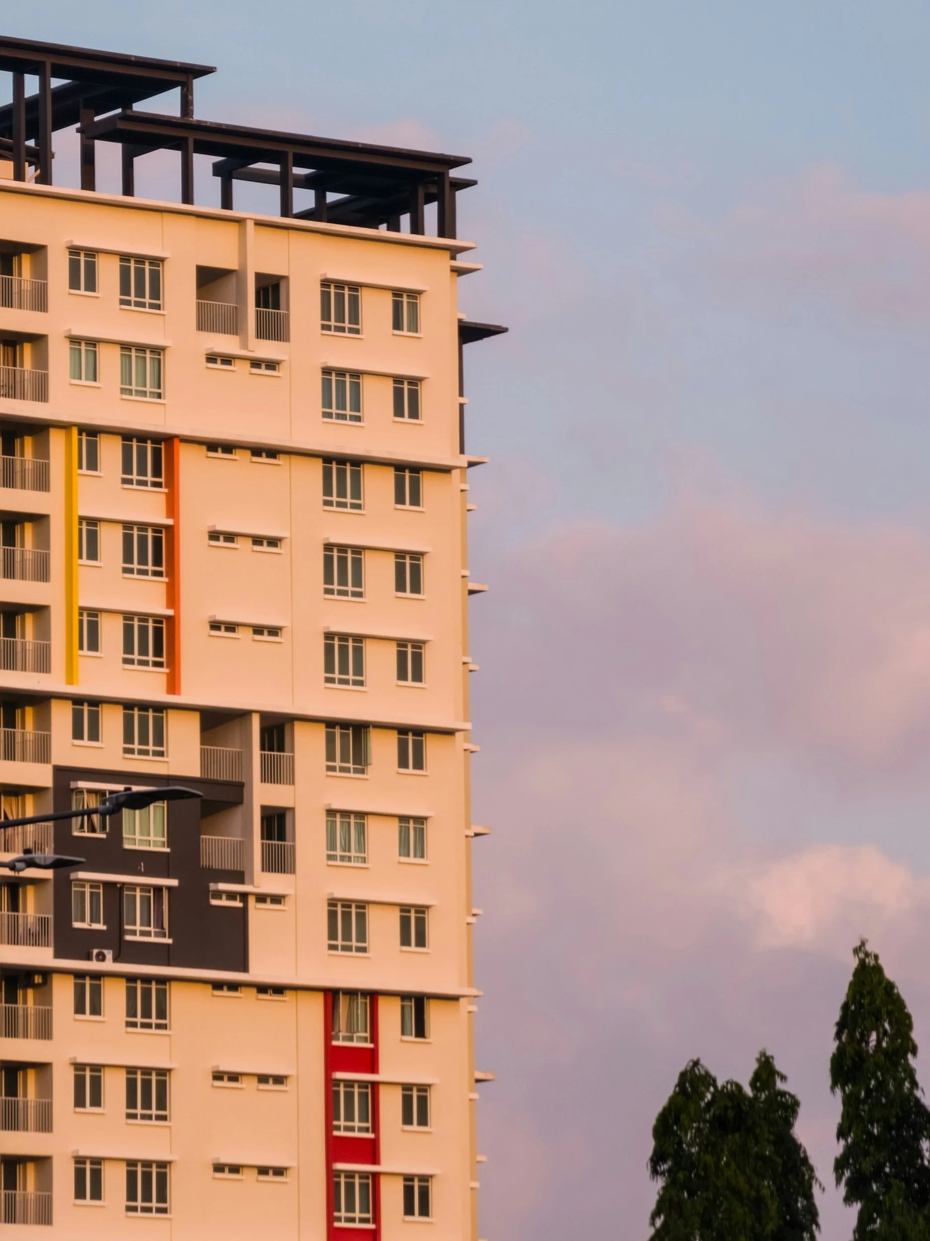 Une photographie d'un immeuble résidentiel moderne avec plusieurs fenêtres, en partie coloré avec des lignes orange, jaune, noire et rouge, sous un ciel avec des nuages et quelques arbres en premier plan.