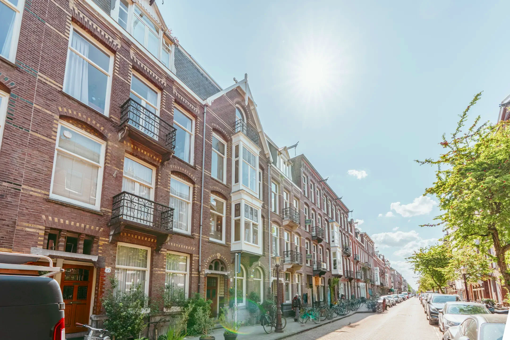 Een straat met historische gebouwen en winkels, auto's geparkeerd langs de stoep, bomen en fietsen, onder een heldere blauwe lucht met de zon die schijnt.