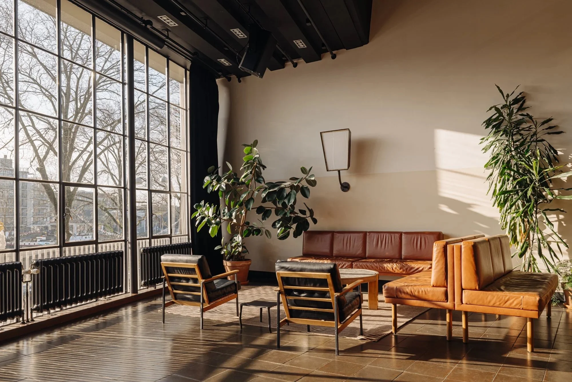 Binnenruimte met grote ramen, een bank, twee chairs, twee planten, en een muurlamp.