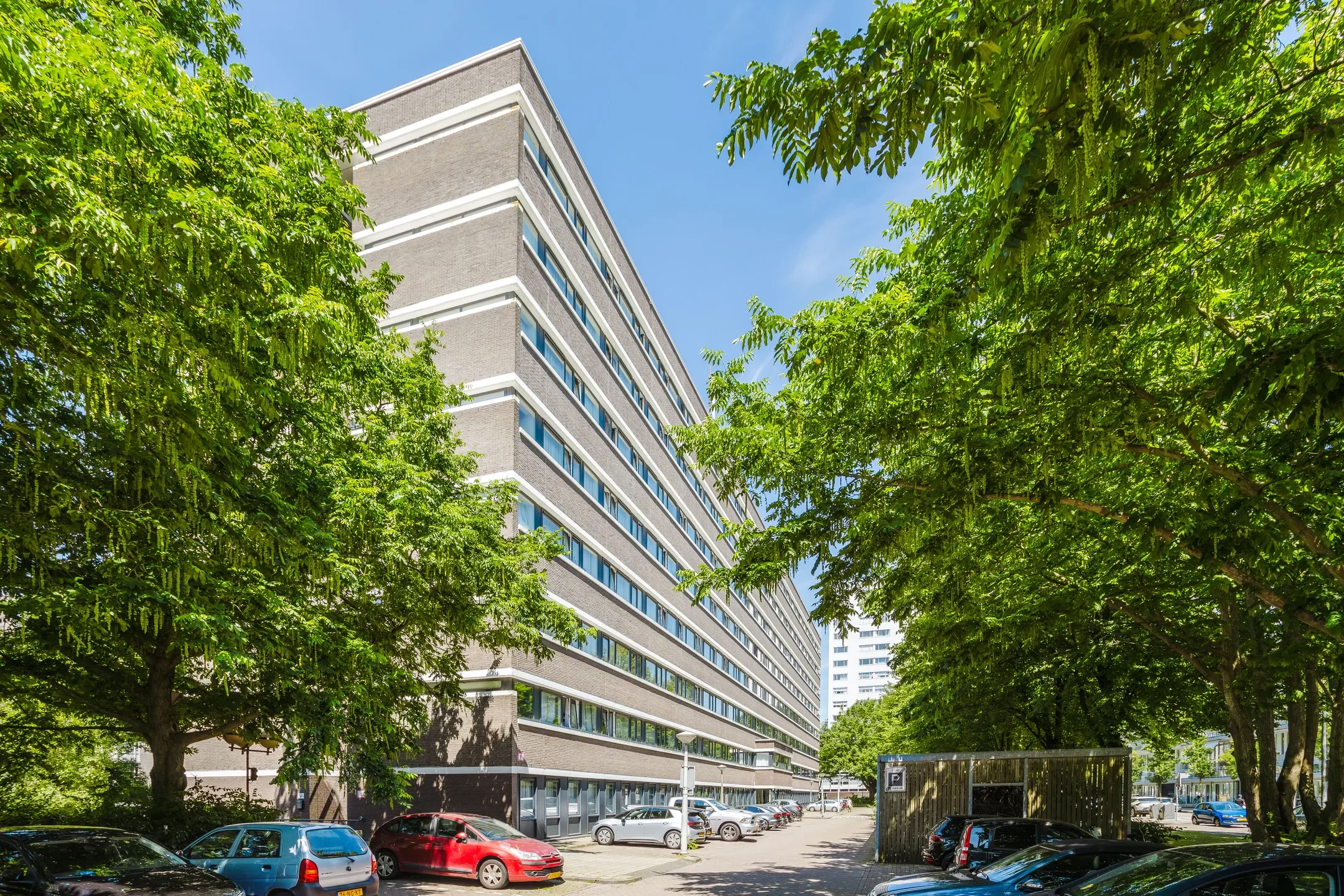 Een modern kantoorgebouw met meerdere verdiepingen, omgeven door groene bomen en een parkeerterrein met auto's, onder een blauwe hemel.