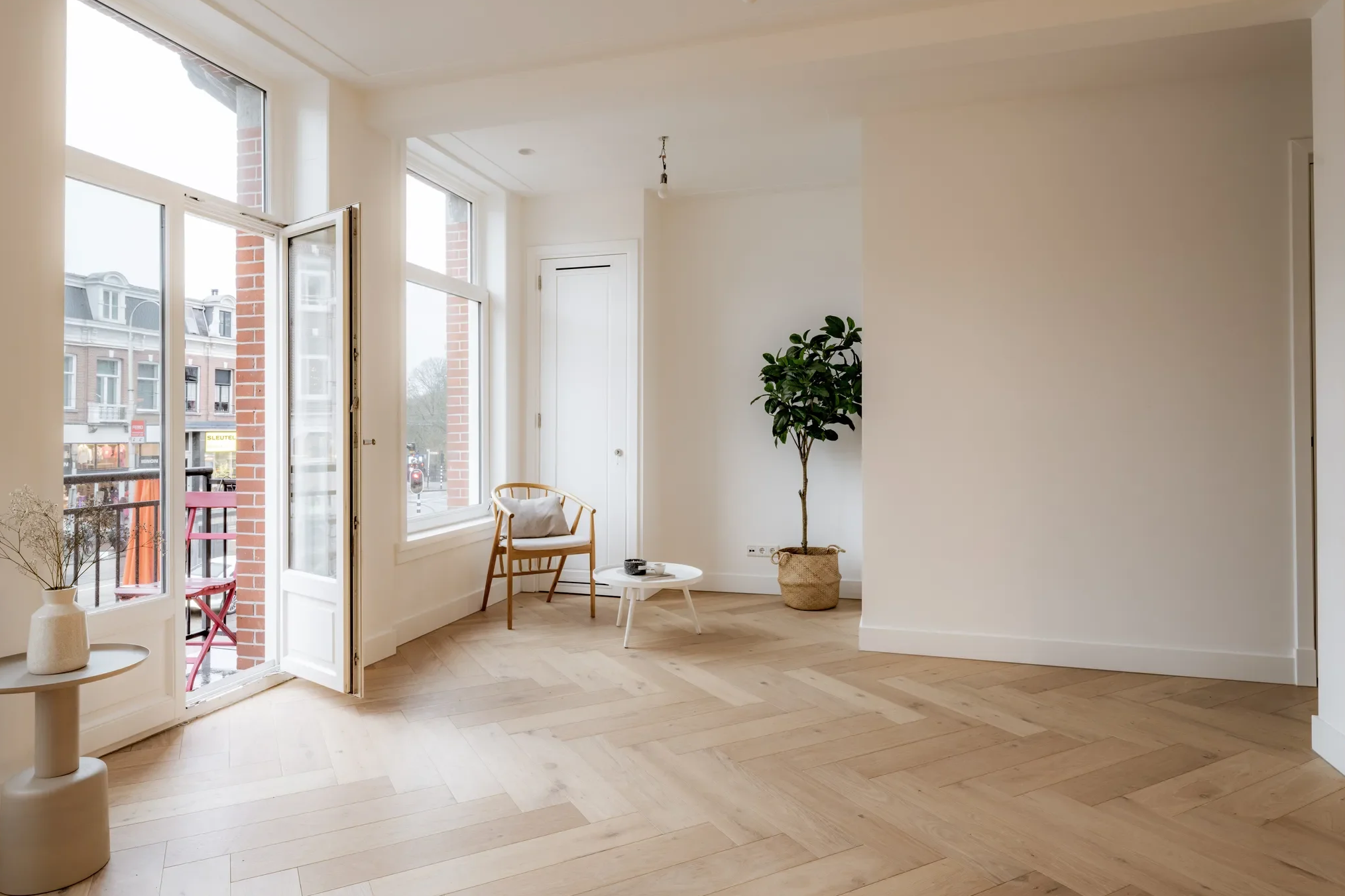Binnenruimte van een modern appartement met houten vloer, grote ramen en een kleine balkon, met een stoel, tafel en een grote plant.