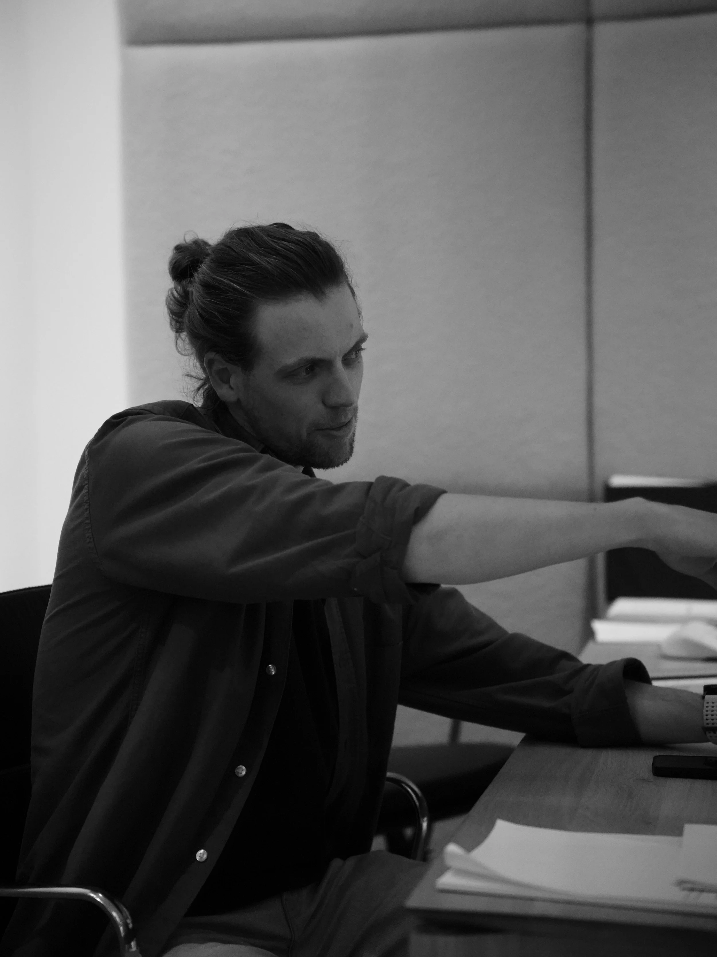 Man met lang haar en een baard zit aan een bureau, hij strekt zijn arm uit en heeft papers voor zich liggen.