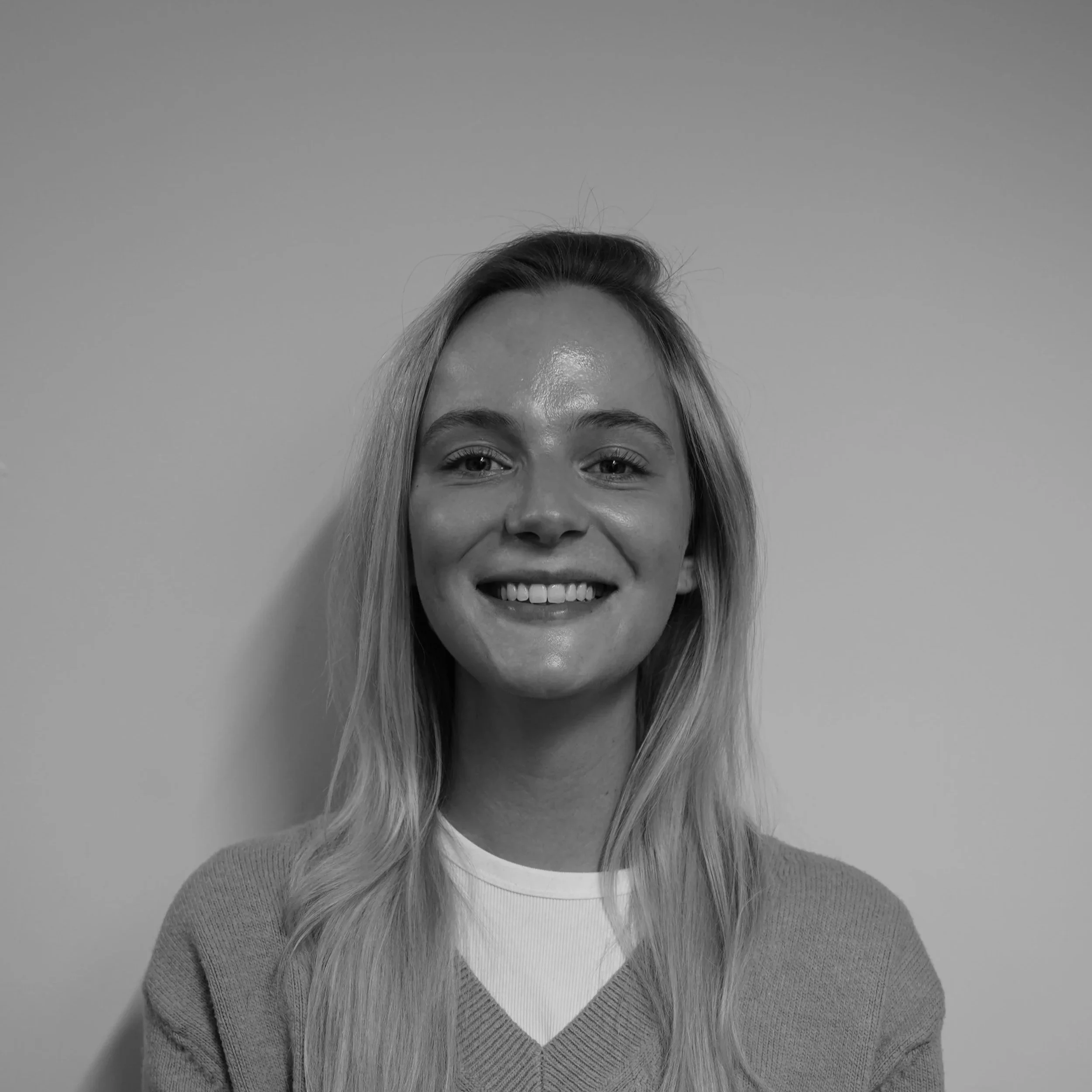 Een zwart-wit foto van een lachende jonge vrouw met lang, licht haar, gekleed in een lichte trui, staand voor een effen muur.