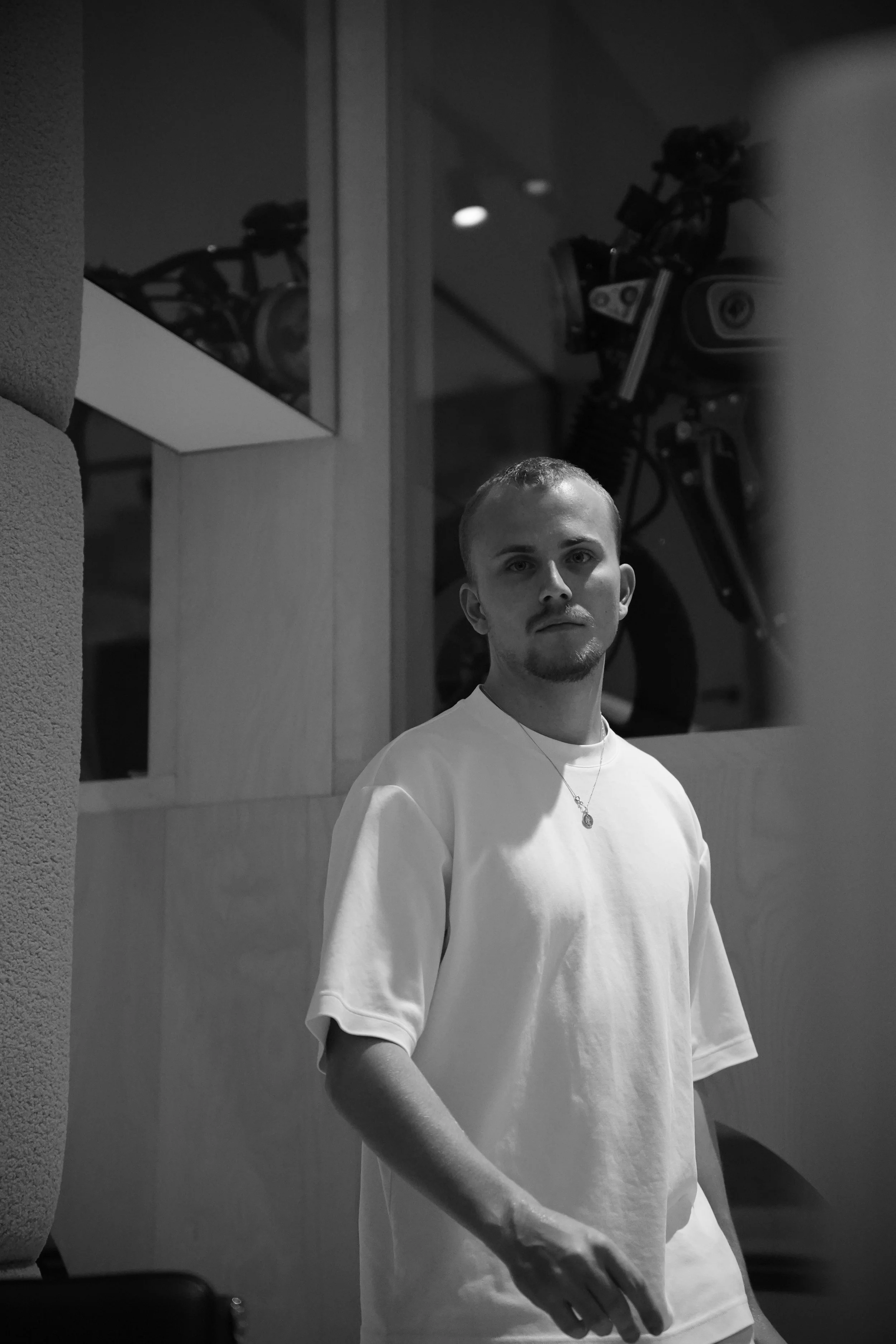 Een jongeman met een lichte huid en kort haar staat binnen, gekleed in een witte T-shirt en een ketting. Op de achtergrond is een motorfiets zichtbaar, en de foto is in zwart-wit.
