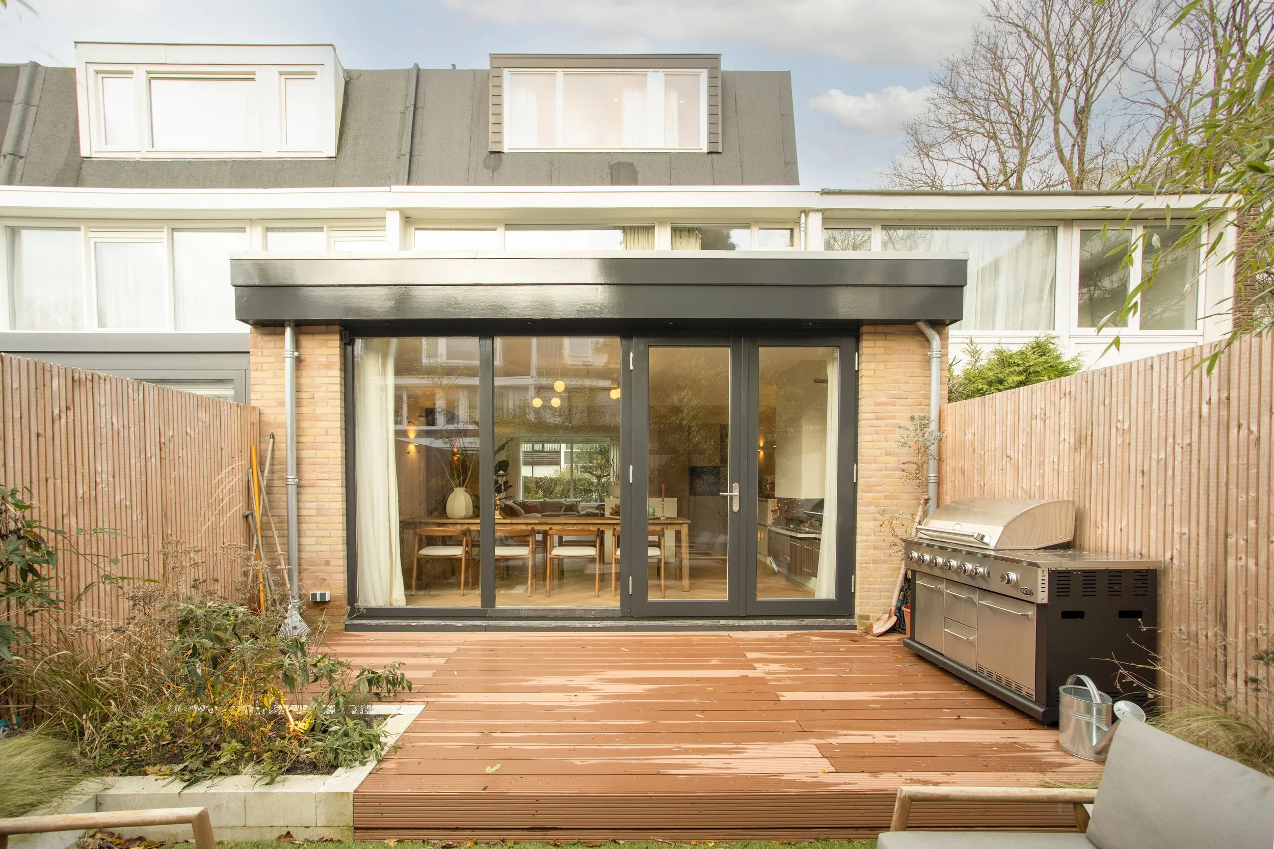 Een patio met houten vloer, een barbecuetoestel en een tuin met planten, achter een huis met grote glazen deuren die naar een woonruimte leiden.