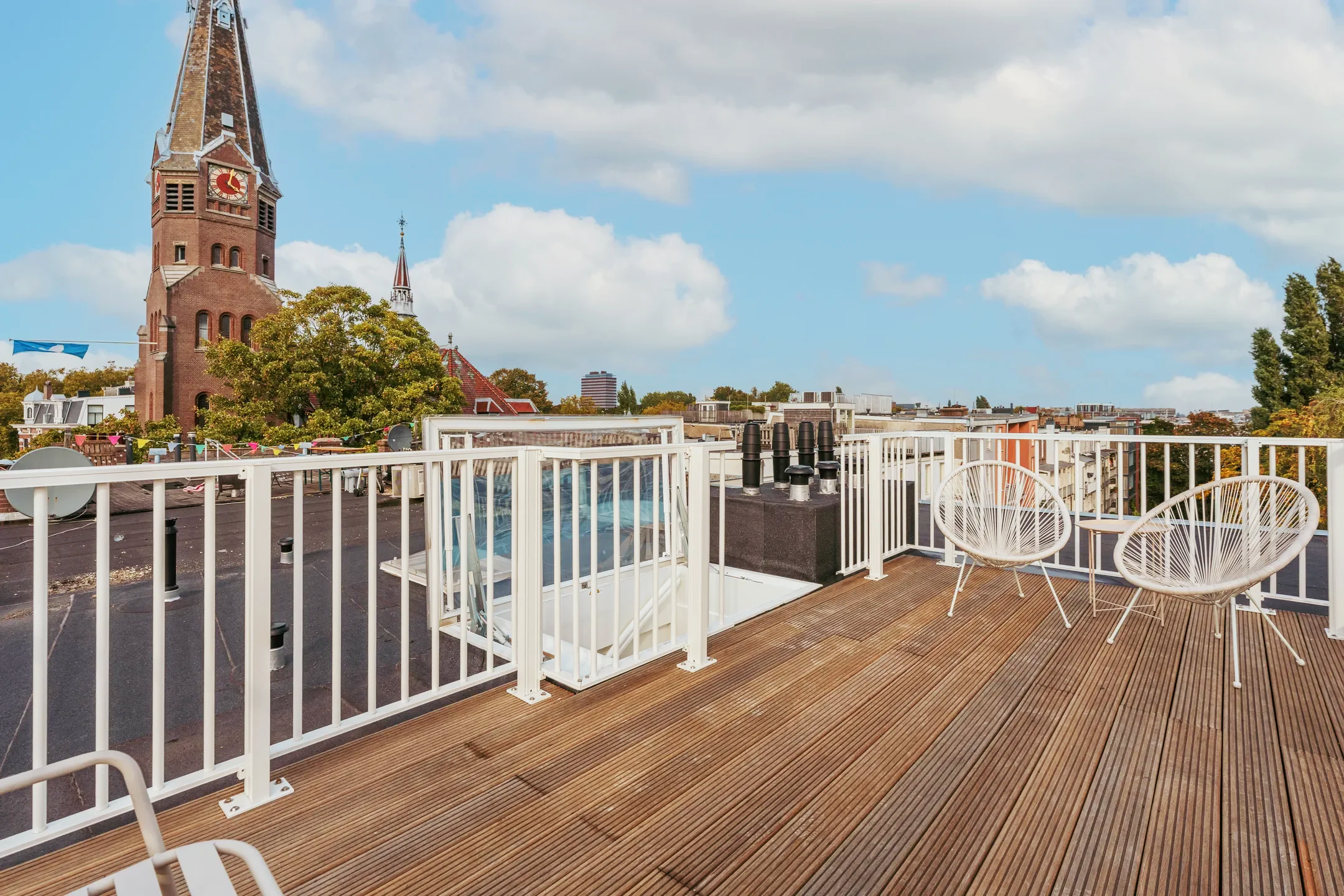 Uitzicht vanaf een dakterras met houten vloer, witte meubels en een kerk met toren op de achtergrond onder een bewolkte blauwe hemel.