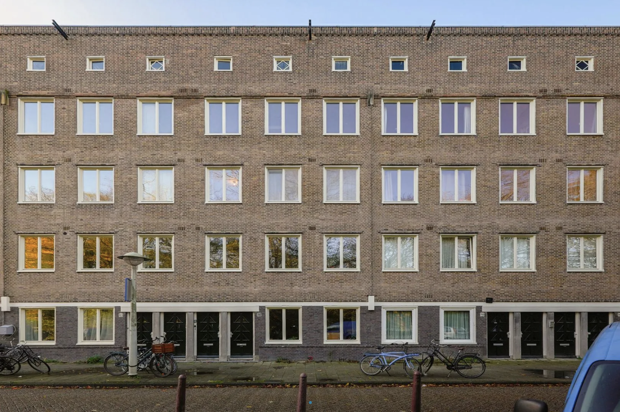 Een afbeelding van een bricken appartementencomplex met meerdere verdiepingen, fietsers voor de ingang en een straat met geparkeerde auto's.