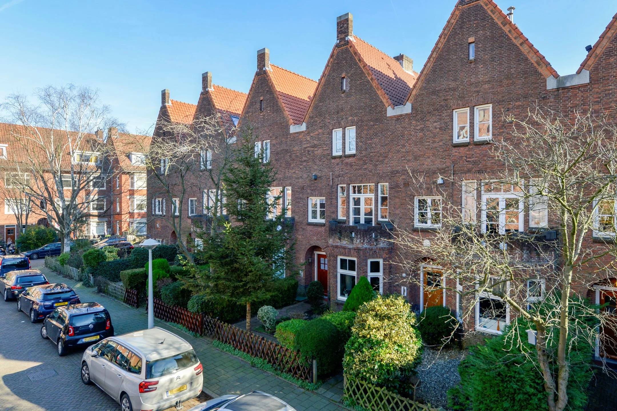 Rij van traditionele Nederlandse bakstenen huizen met rood recht dak, bomen en geparkeerde auto's langs de straat op een heldere dag.