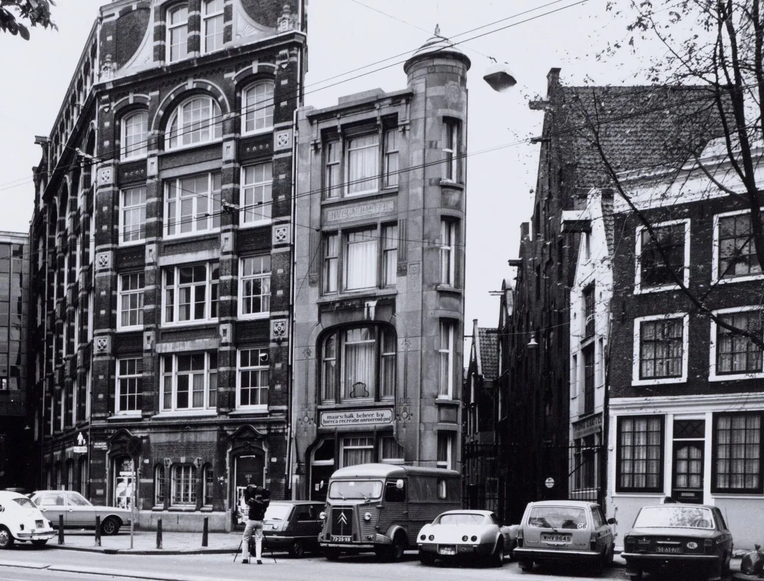 Een gedrongen geel/bruine hoekwoning met meerdere verdiepingen en smalle ramen, naast andere gebouwen op een stadspand. Borden bij de ingang en fietsen voor de deur.