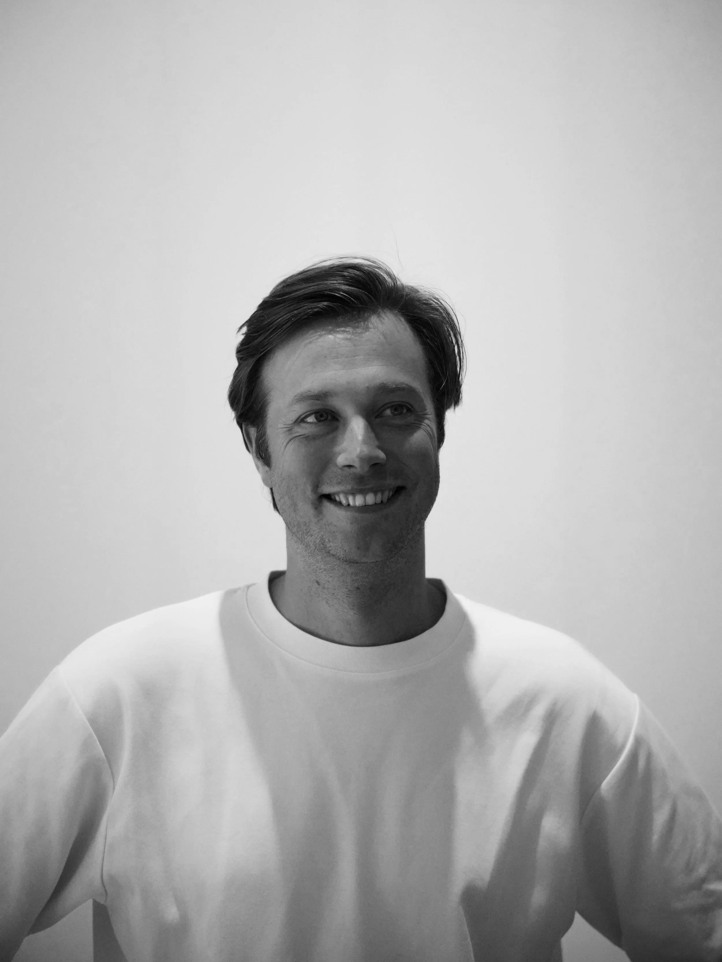 Zwart-wit foto van een glimlachende man met halflang haar, gekleed in een witte T-shirt, staand voor een witte muur.