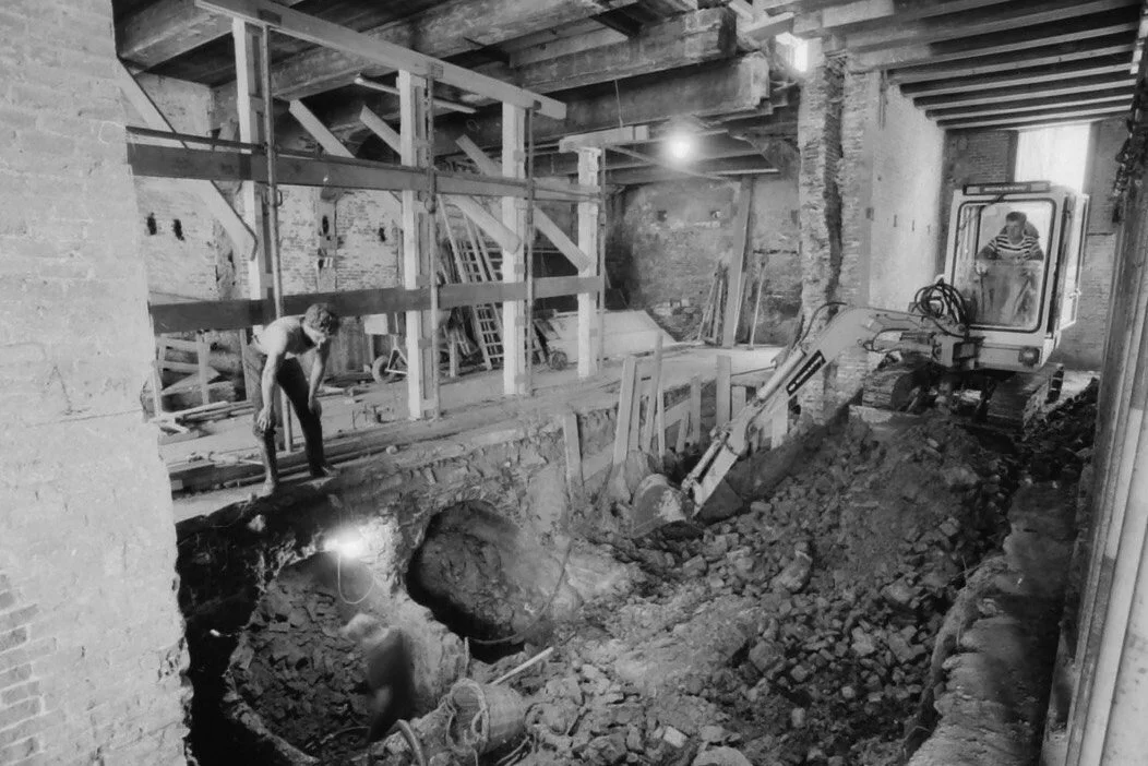 Binnen een bouwplaats met een graafmachine en enkele bouwvakkers die werken aan de fundering of de aanwezige structuur van een gebouw, met bakstenen muren en houten steunen.