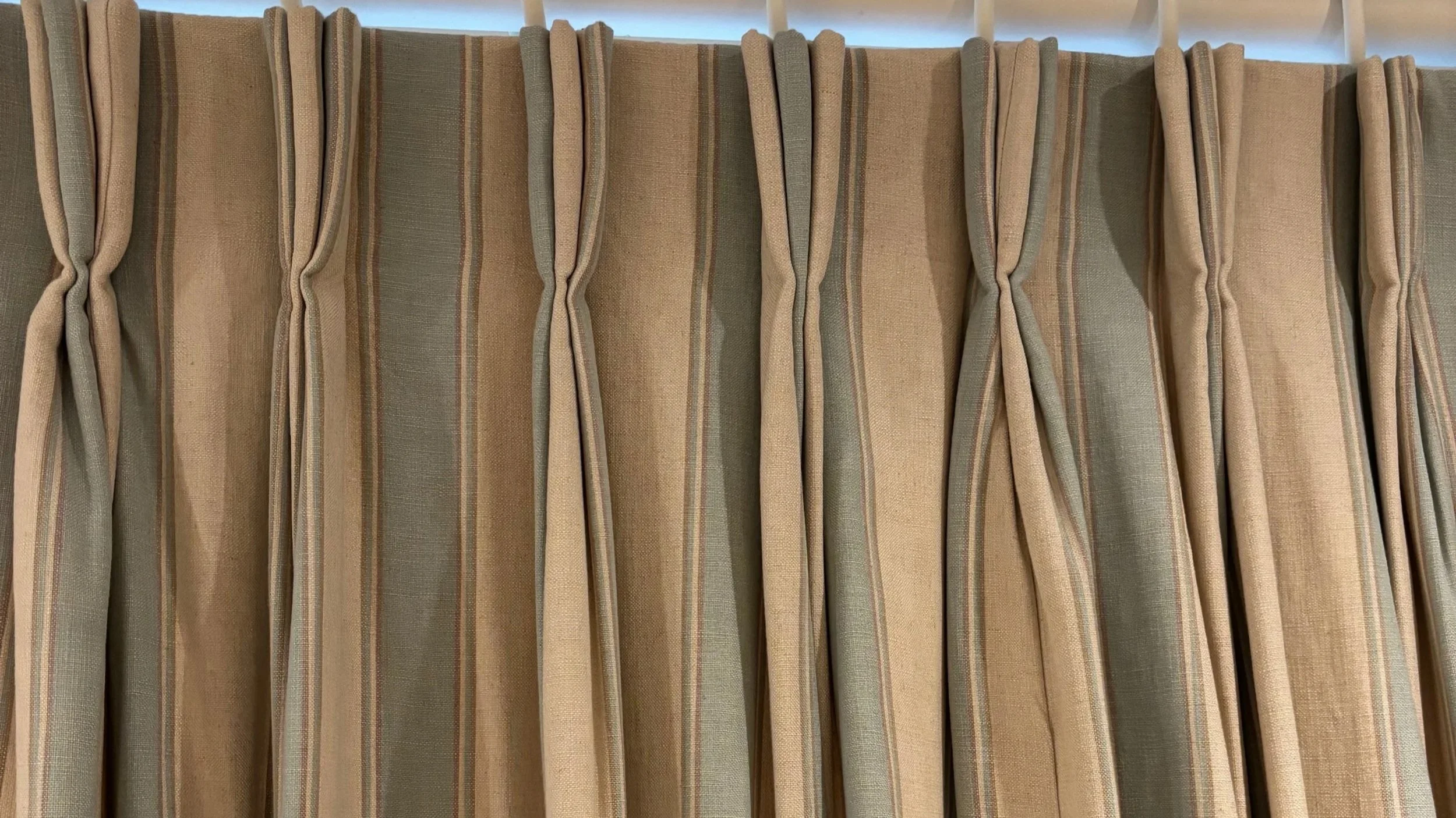 Beige-striped pleated curtains hanging from a curtain pole indoors.
