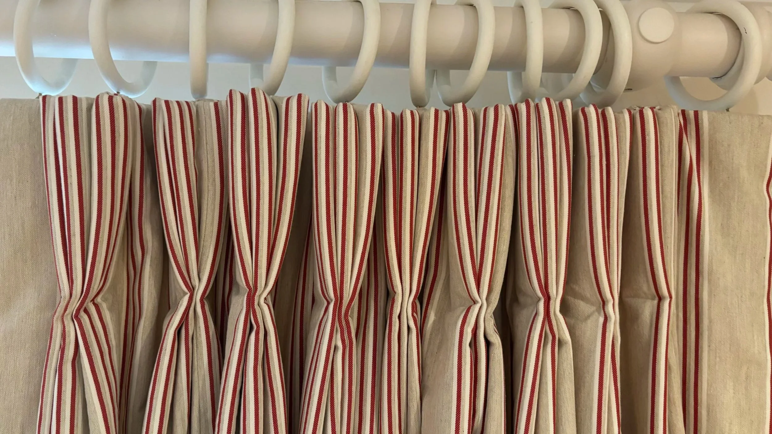 Beige curtains with red and white striped fabric along the top, hanging from a white wooden curtain pole with multiple rings.