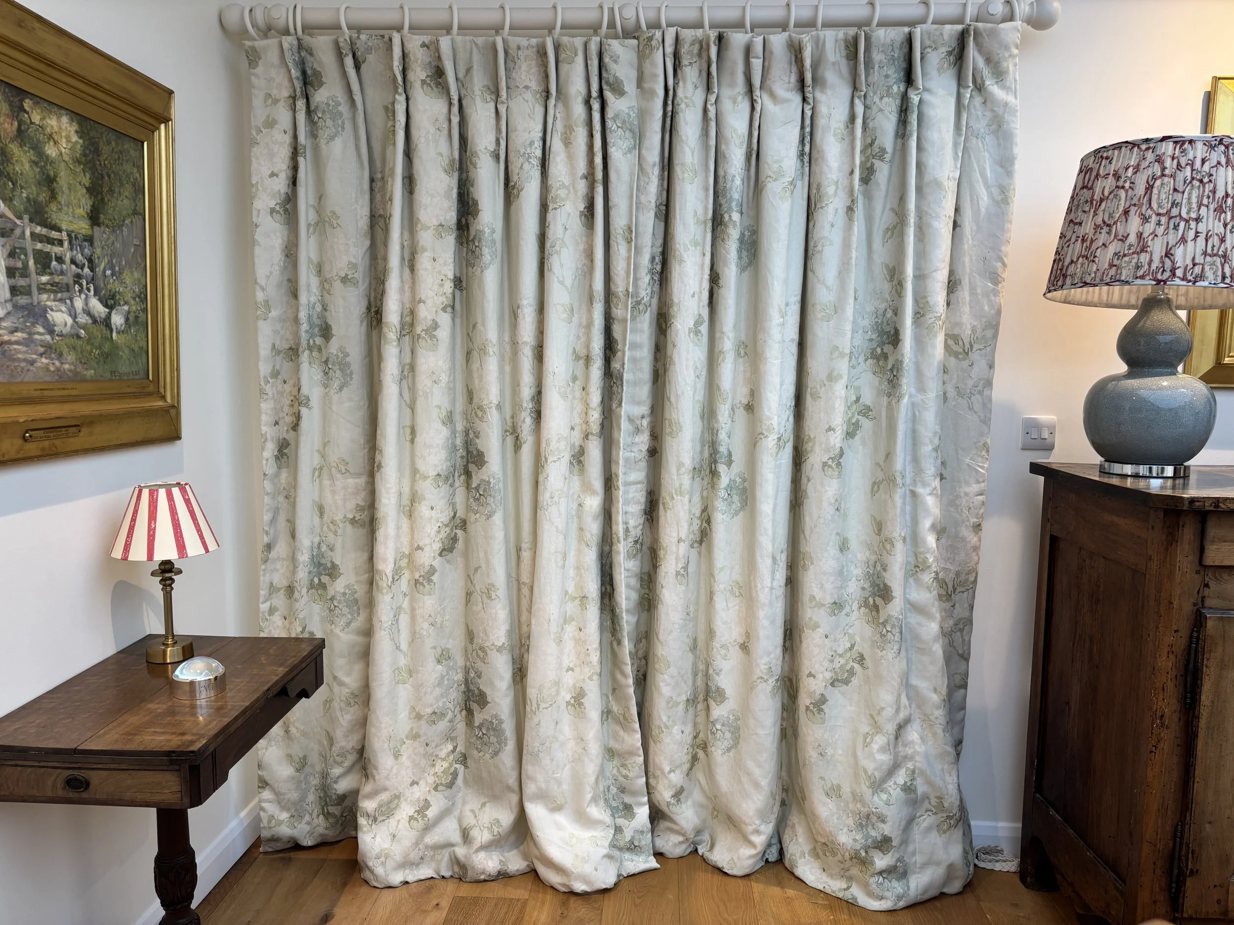 Indoors, beige floral curtains covering a window, flanked by a wooden side table with a small striped red and white lampshade and a decorative item, and a tall wooden cabinet with a large gray ceramic lamp with a patterned shade.