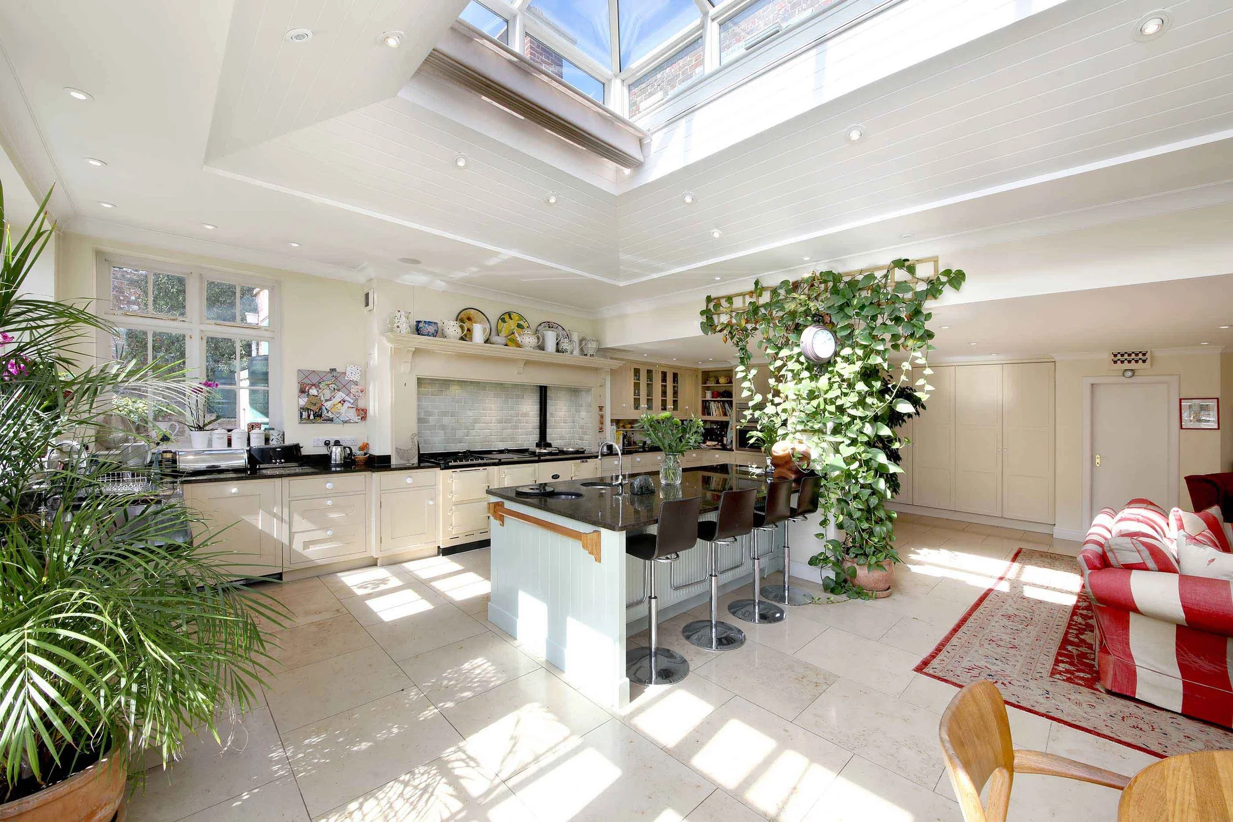 Bright, spacious kitchen with an open layout, skylight ceiling, white cabinetry, black countertops, a central island with barstools, potted plants, and a seating area with a red and white striped sofa.