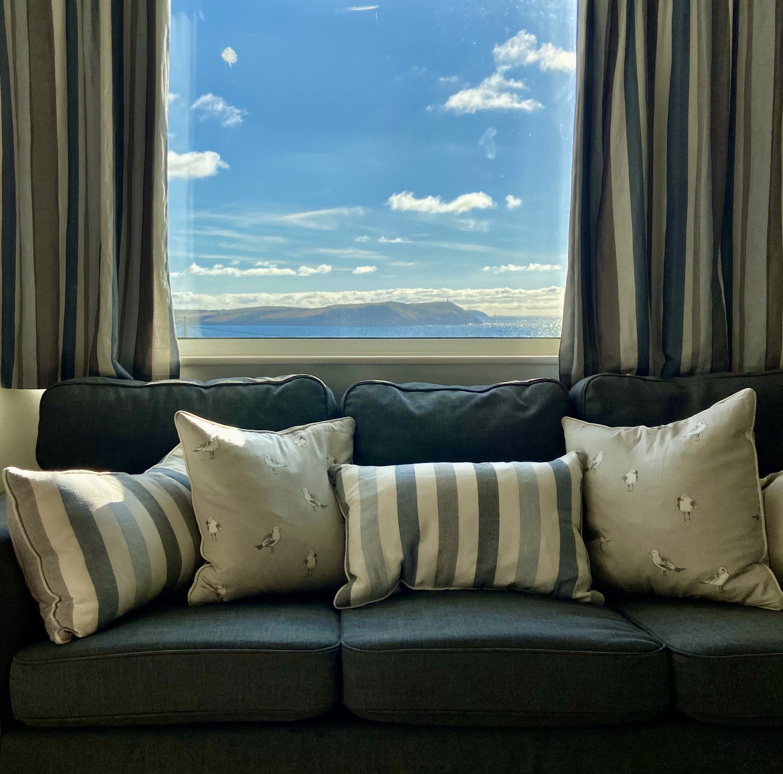 Living room with a dark blue sofa and patterned pillows, large window with curtains, blue sky with clouds and a distant landscape visible through the window.