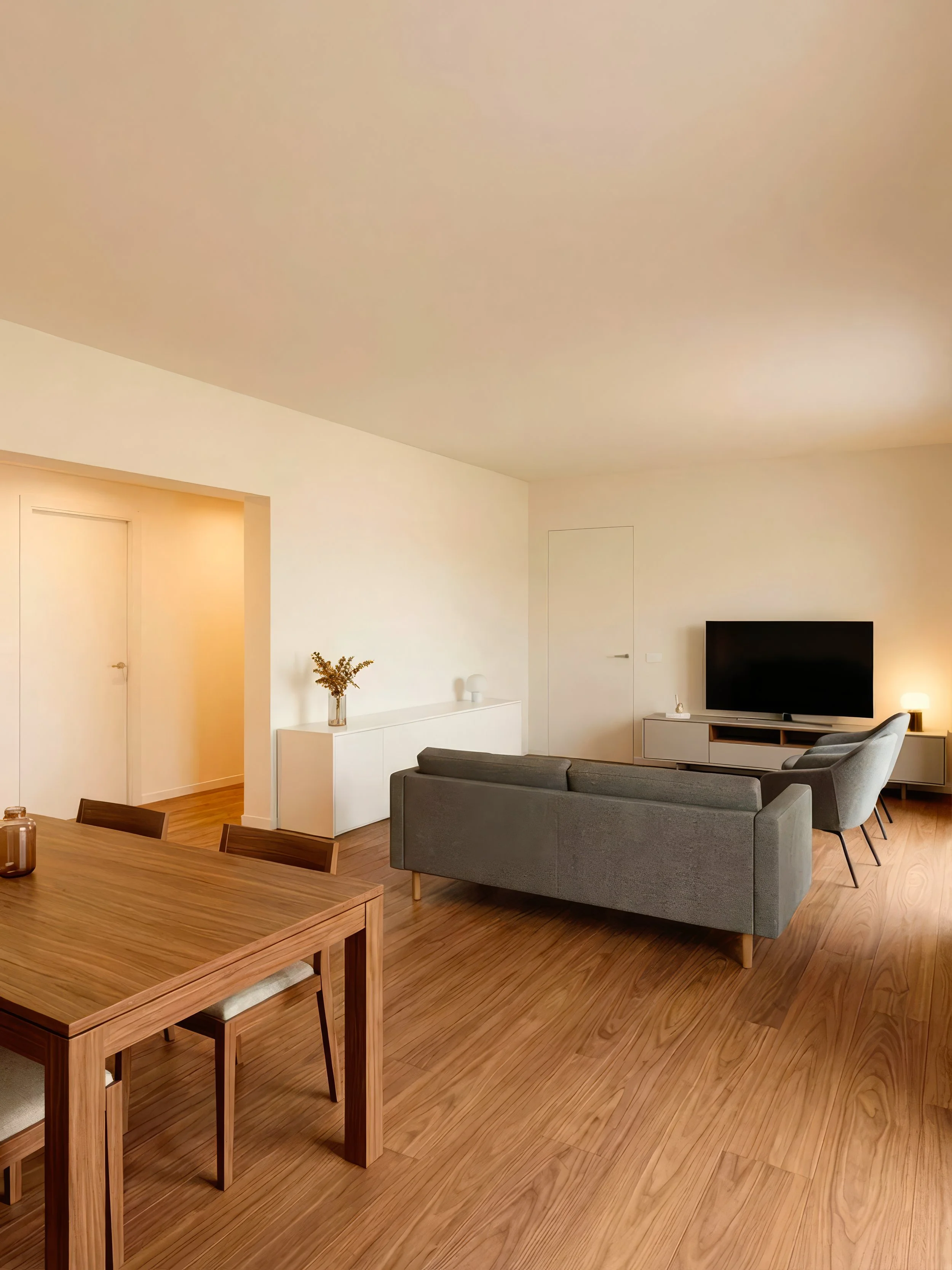 Salotto con divano grigio, due poltrone grigie, TV su mobile bianco, lampada accesa, tavolo in legno con sedie, pareti bianche, pavimento in legno.