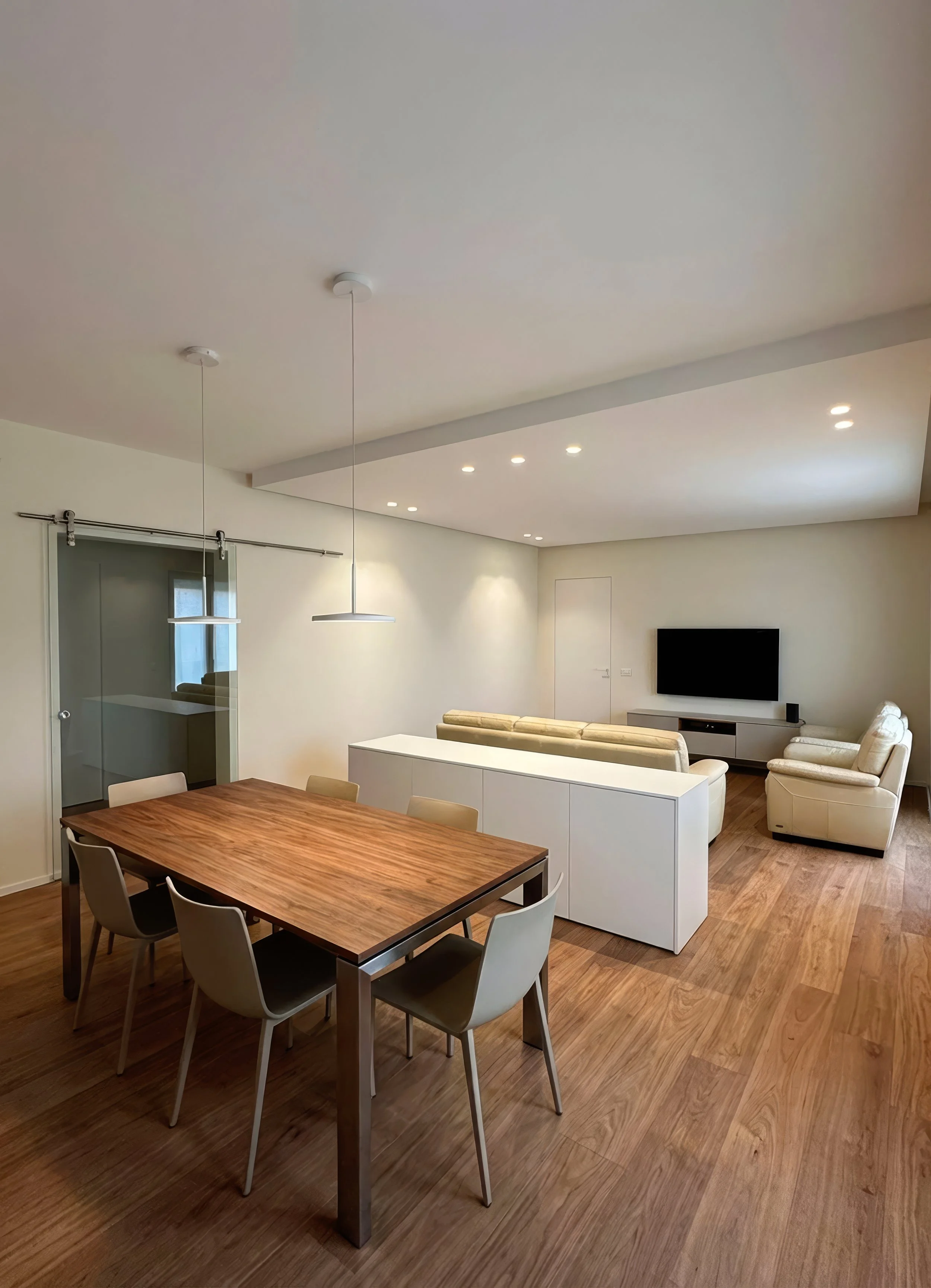 Soggiorno con tavolo da pranzo in legno, sedie beige, divani bianchi, TV a parete, soffitto con luci a incasso, pareti bianche, pavimento in legno.