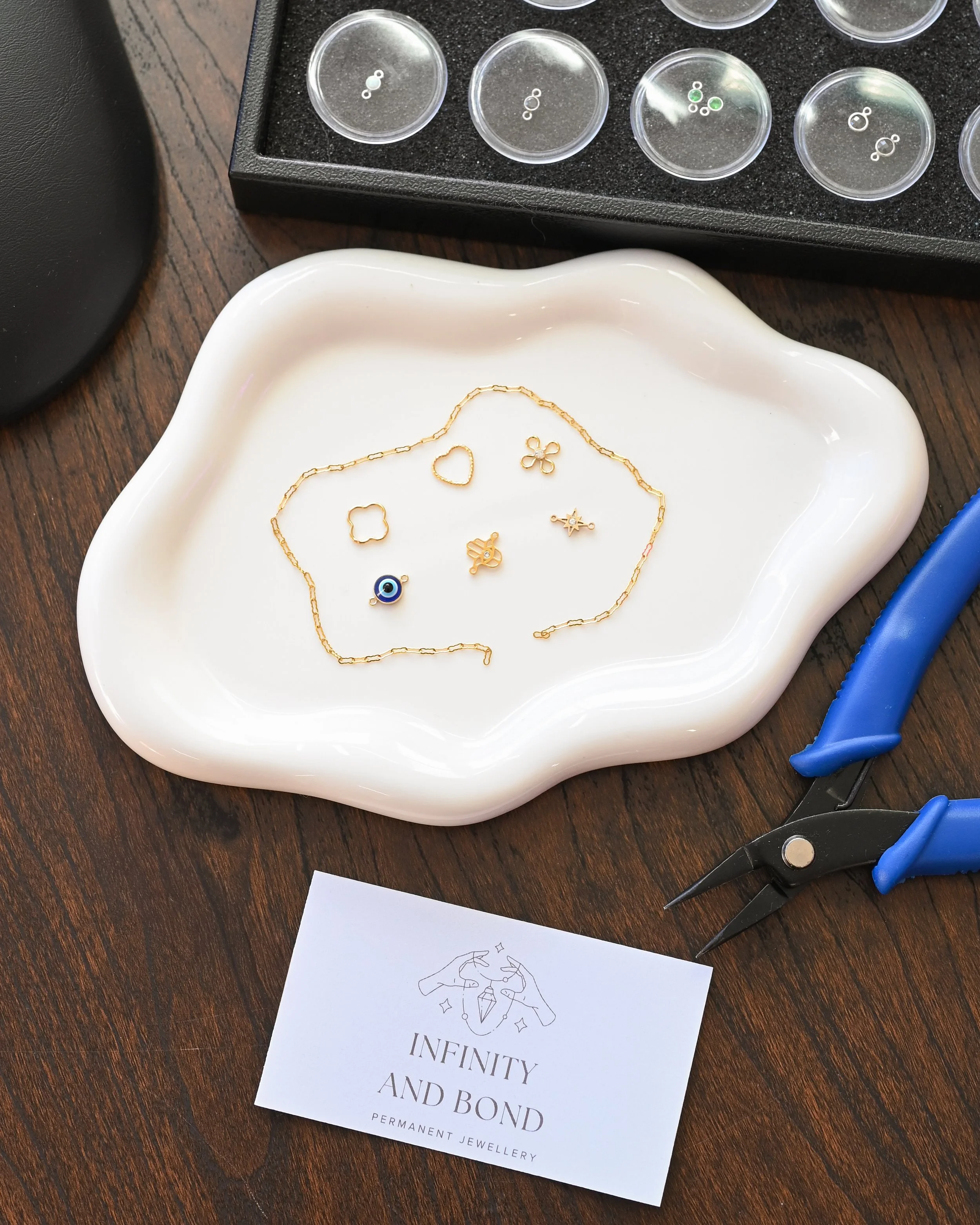 A white ceramic dish containing gold jewelry such as delicate chain necklaces and small charms, gardening tools like pliers, a card with the logo and text 'Infinity and Bond Permanent Jewellery', and a black box with small clear containers of tiny beads or charms.