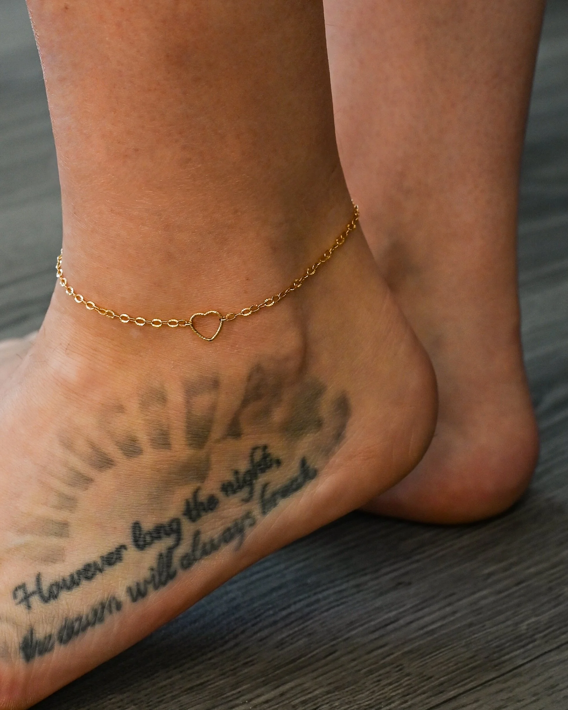 Close-up of a person's ankle with a gold chain anklet featuring a heart shape, and a tattoo of a quote on the forearm.
