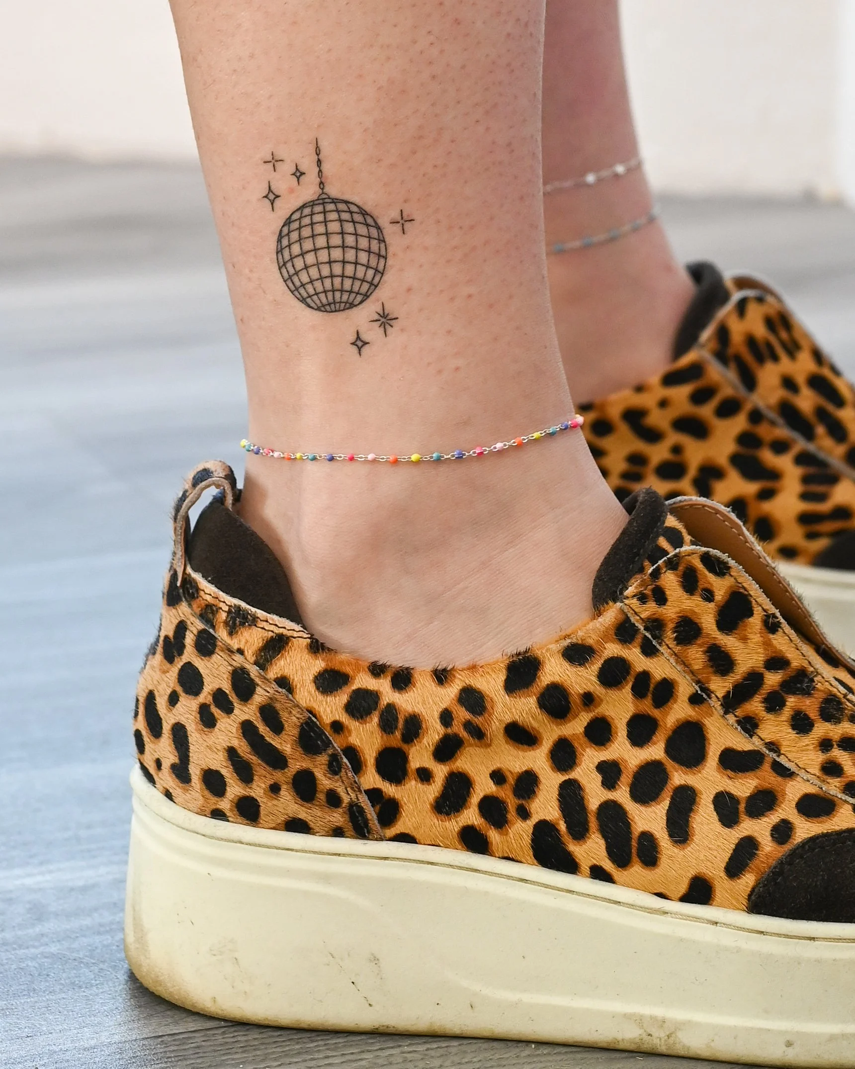 Close-up of a person's ankle showing a small colorful beaded anklet, a tattoo of a disco ball with stars, wearing leopard print sneakers on a wooden floor.