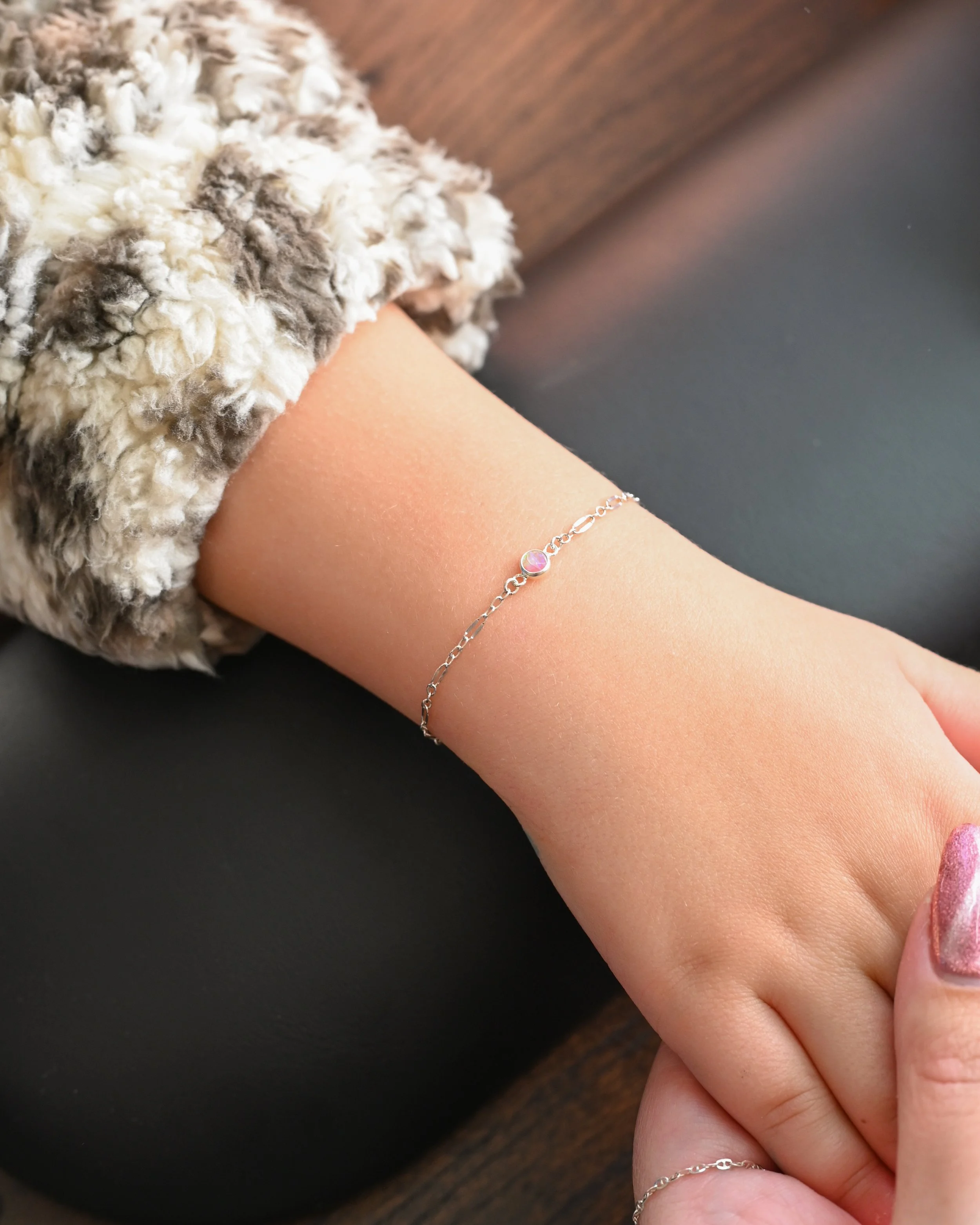 A person's wrist with a delicate silver bracelet featuring a small pink bead, resting on a dark surface and partially covered with a fluffy, textured, beige and gray sleeve.