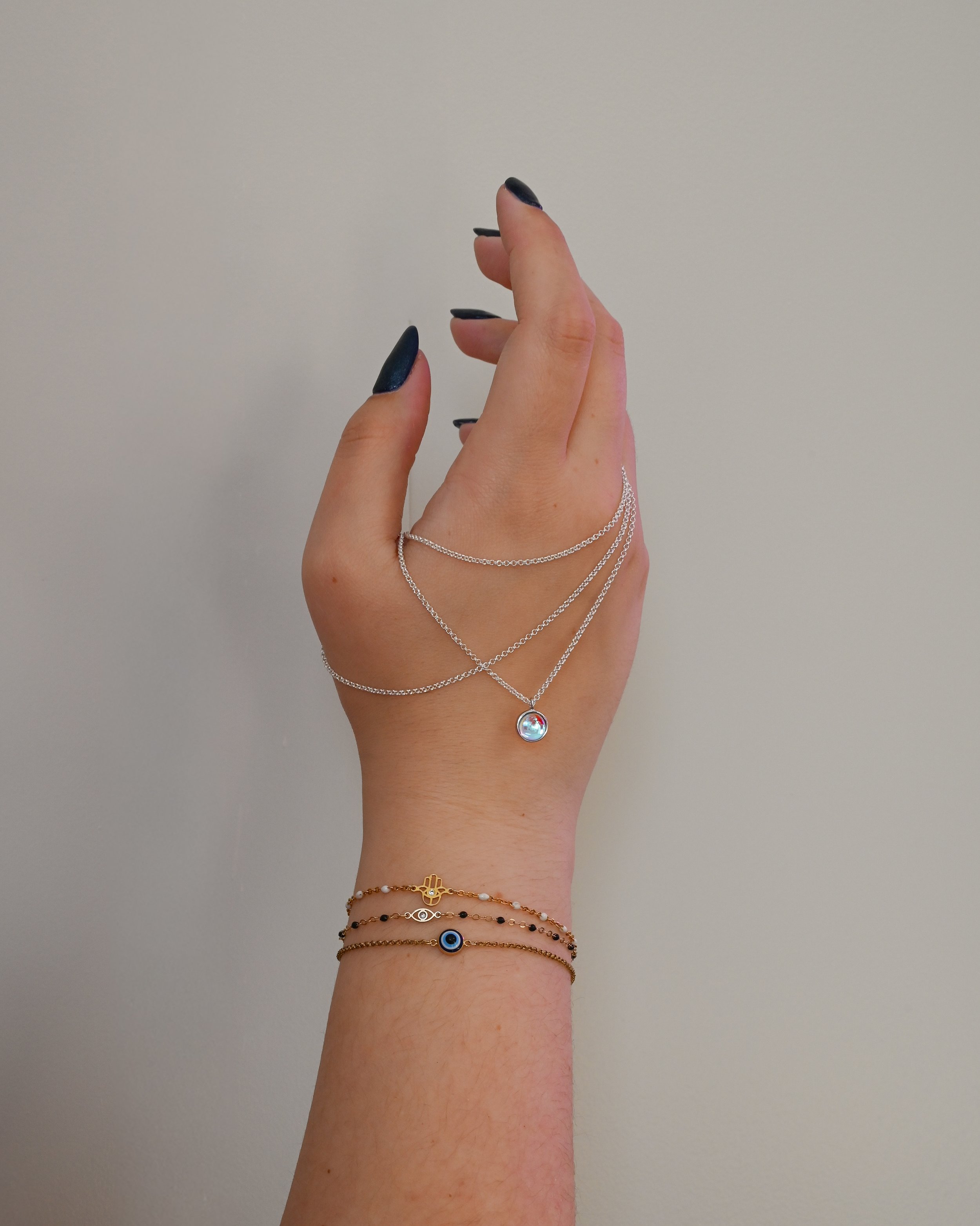 A woman's hand with black painted nails wearing layered silver necklaces and gold bracelets with black, white, and blue beads, and a hamsa charm, against a plain beige background.