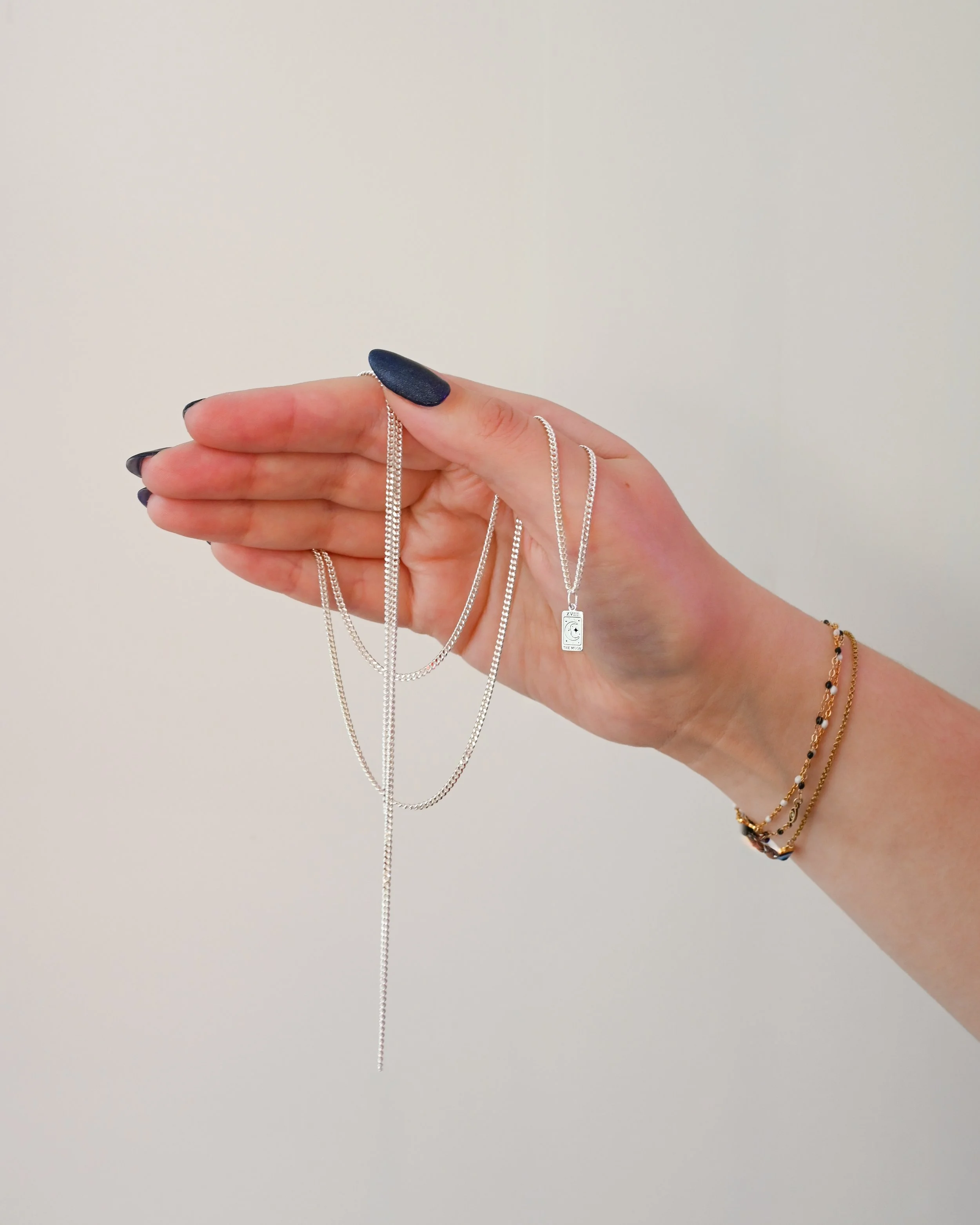 Hand with dark nail polish holding multiple silver necklaces, including one with a pendant, against a plain white background, and wearing layered gold bracelets.