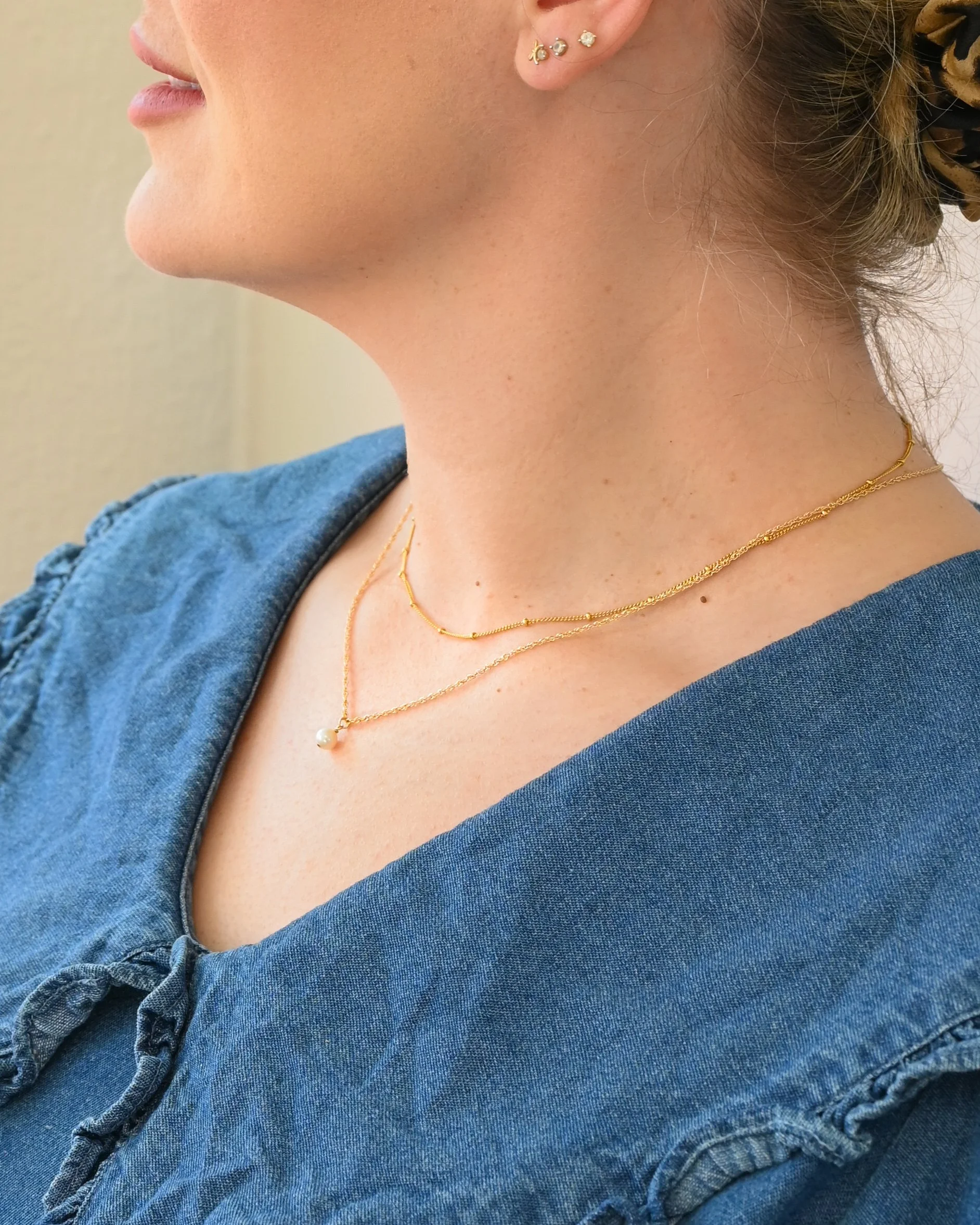 Close-up of a woman's neck and lower face, wearing layered gold necklaces and small gold earrings, with her hair tied back with a leopard print scarf, and dressed in a blue denim top.