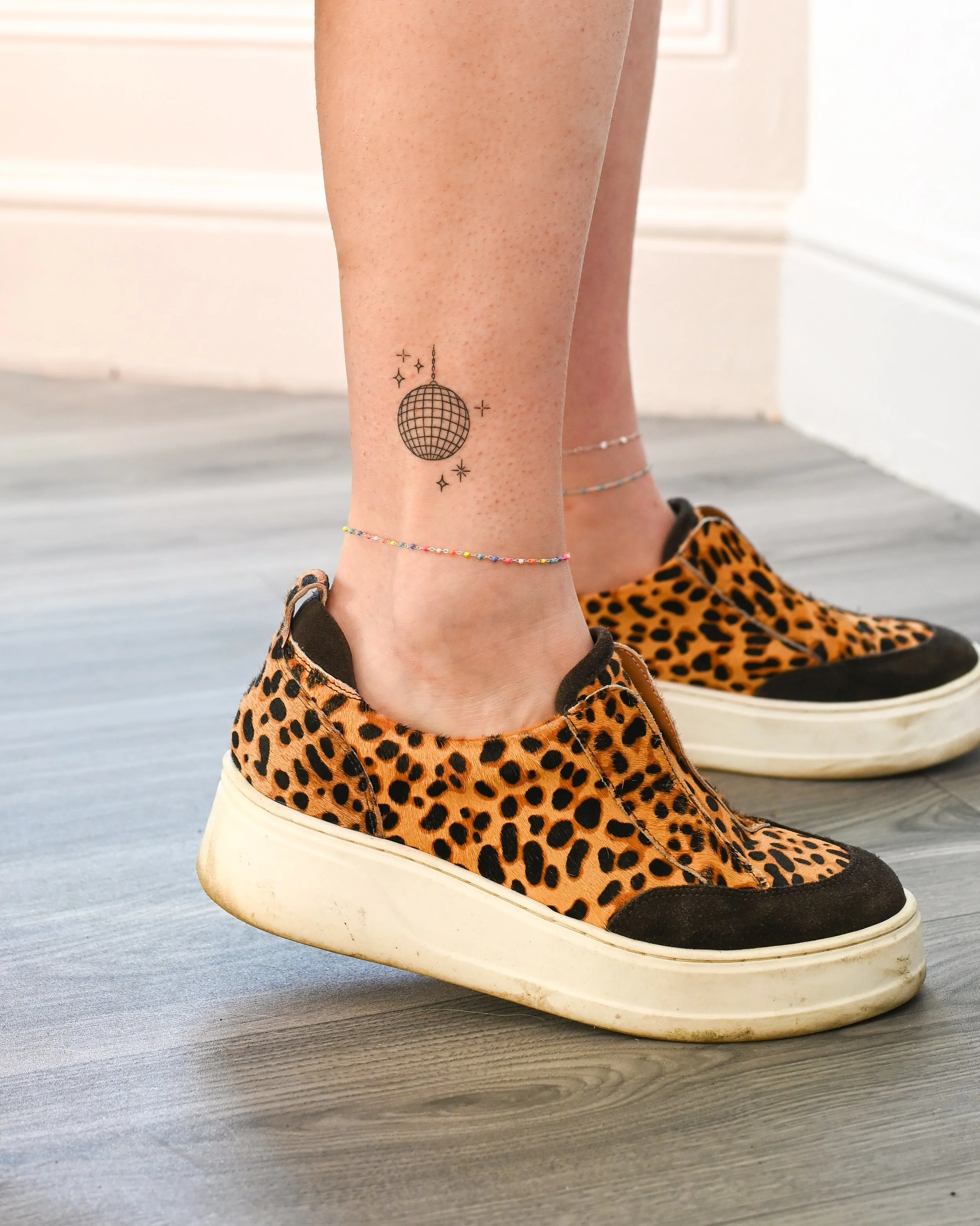 Person wearing leopard print sneakers with black accents, standing on a gray wooden floor, with a tattoo of a disco ball and stars on their ankle, and a colorful beaded anklet.