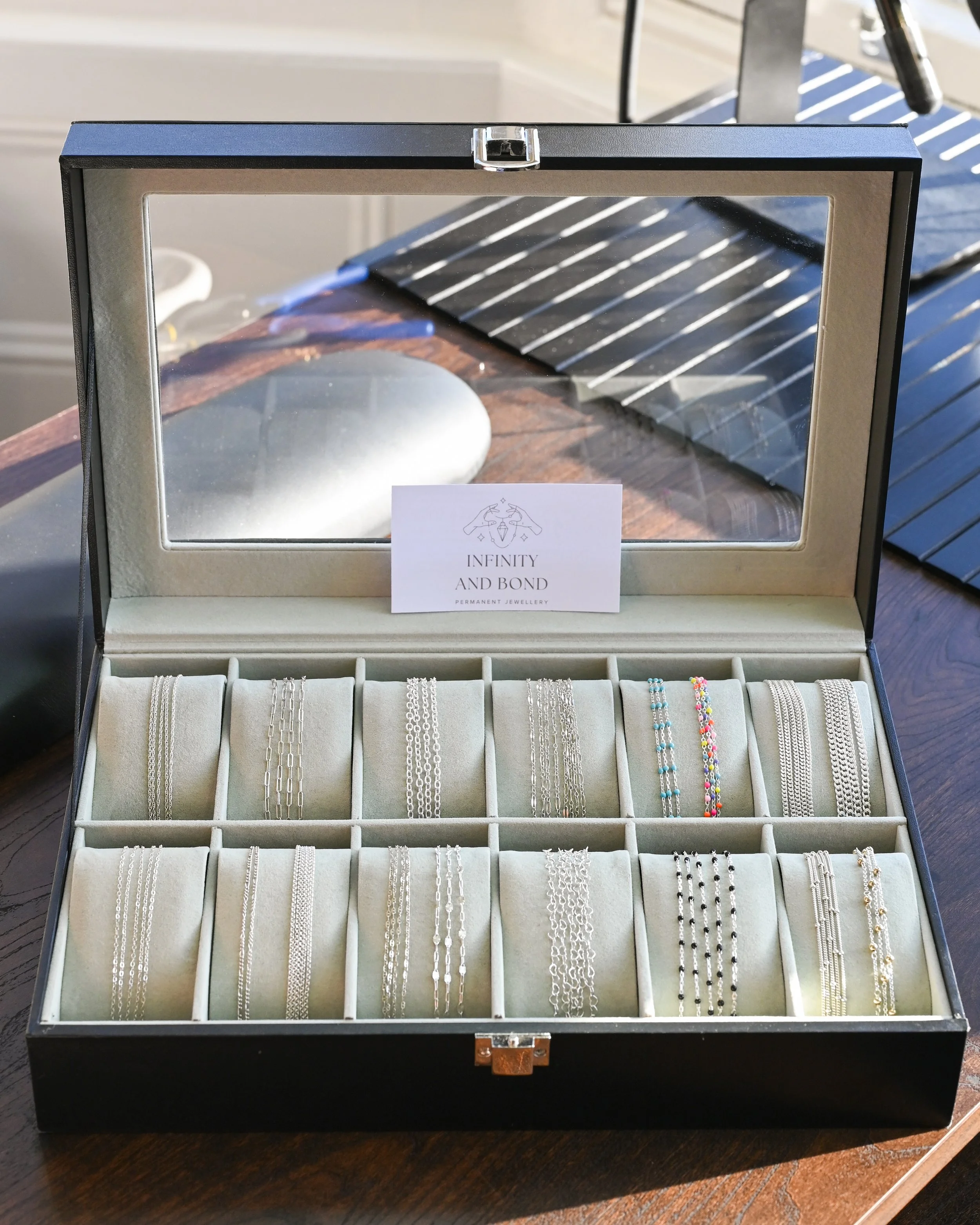 A jewelry showcase box containing multiple silver bracelets and anklets, with a sign reading 'Infinity and Bond' and 'Permanent Jewelry' inside.