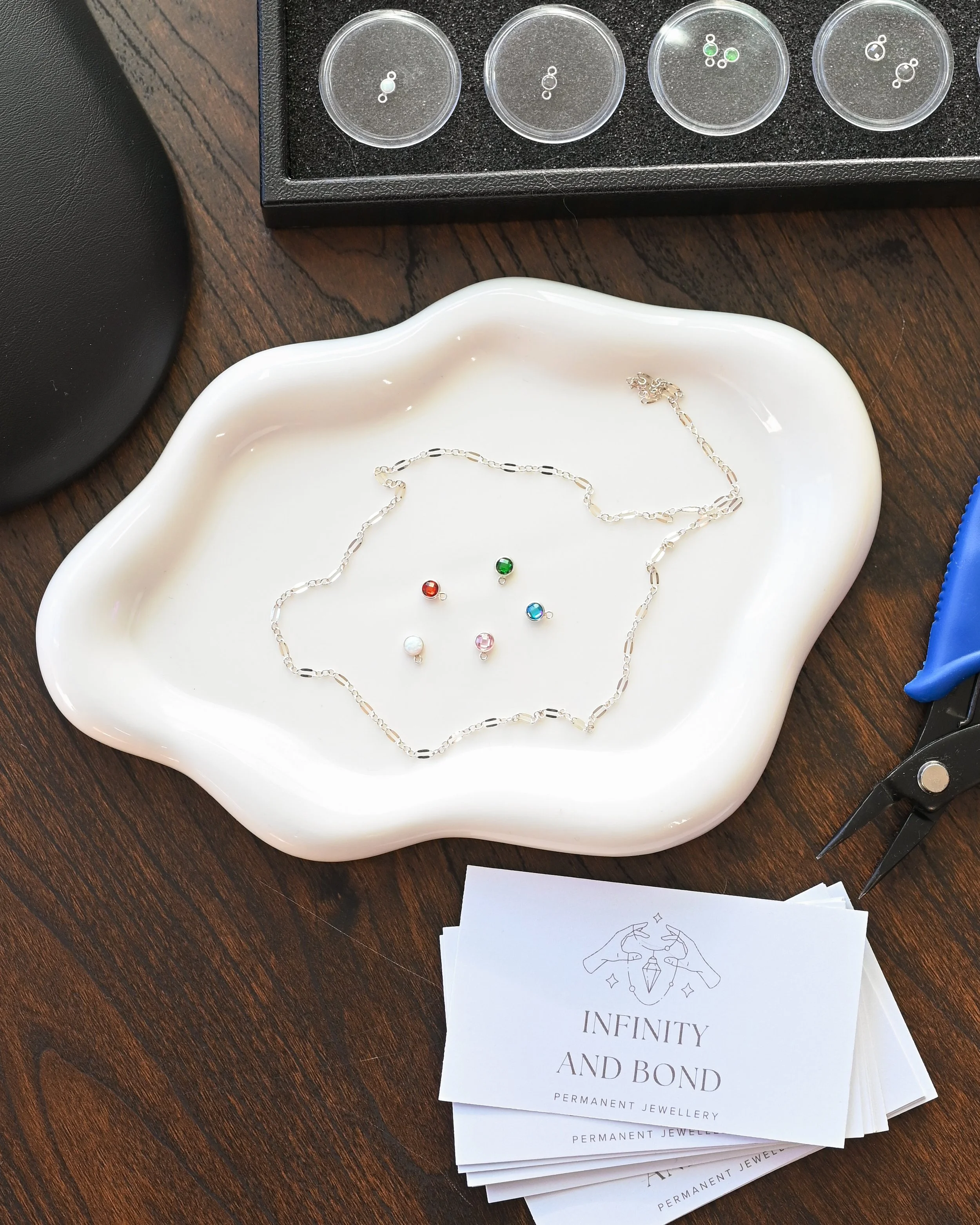 Jewelry-making workspace with a white dish containing a silver chain necklace, five small colored beads, a pair of blue-handled scissors, and a business card for Infinity and Bond permanent jewelry. In the background, there is a black tray with small containers of different colored beads.
