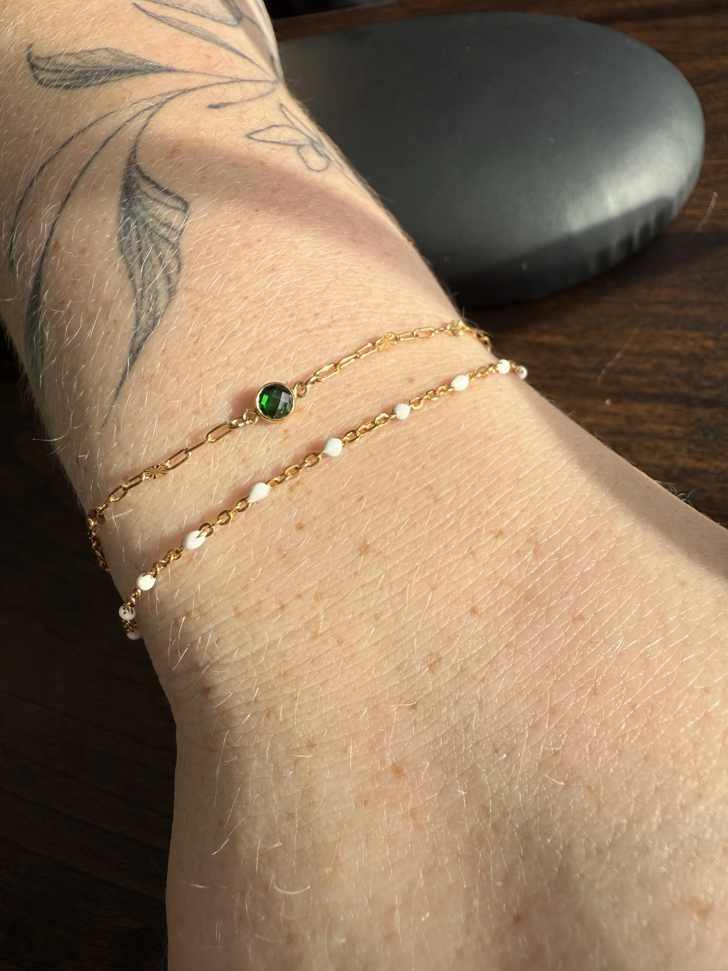 Close-up of a person's wrist with three gold bracelets, one featuring a green gemstone, and a tattoo of a feather on the arm. The background shows a black object and a wooden surface.
