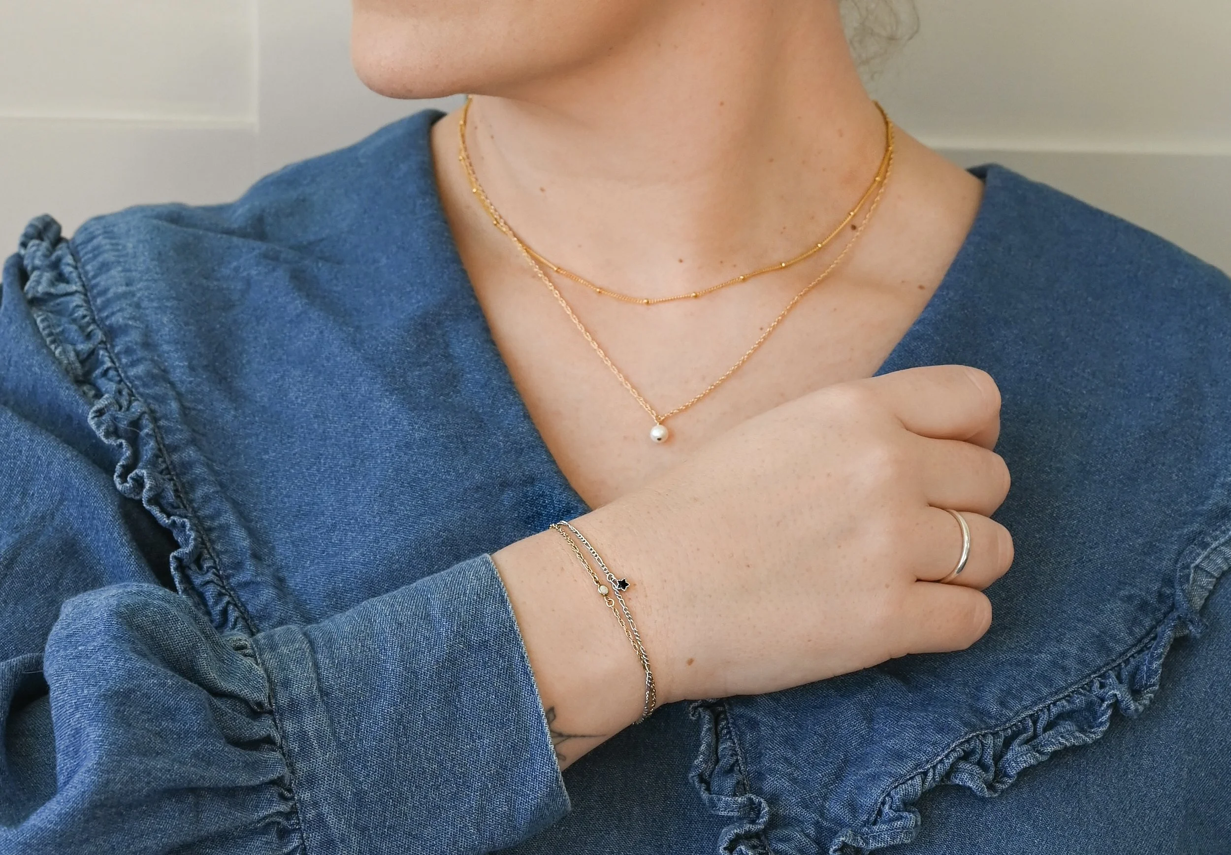 Close-up of a woman wearing layered gold and silver necklaces, a delicate bracelet with a black star and pearl charm, a plain silver ring, and a denim top with ruffled sleeve details.