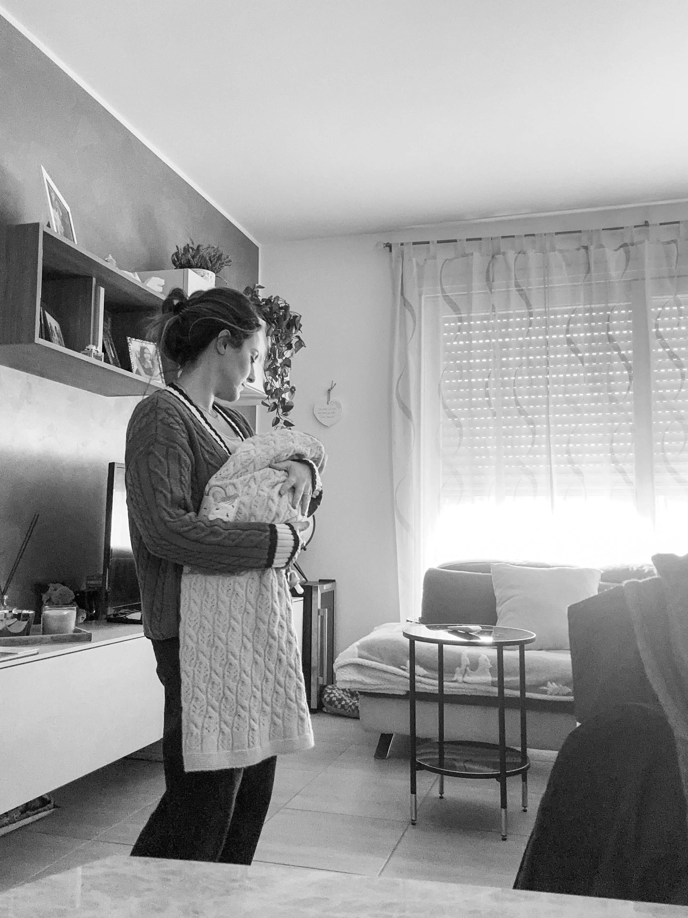 Una donna tiene un neonato avvolto in una coperta nella stanza di casa con finestre, tende e mobili.