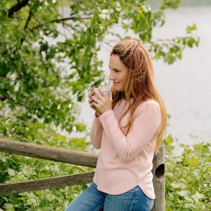 Una donna con lunghi capelli castani tiene una tazza tra le mani, sorridendo e guardando verso destra, all'aperto vicino a un lago e circondata da vegetazione verde, con un molo in legno vicino a lei.