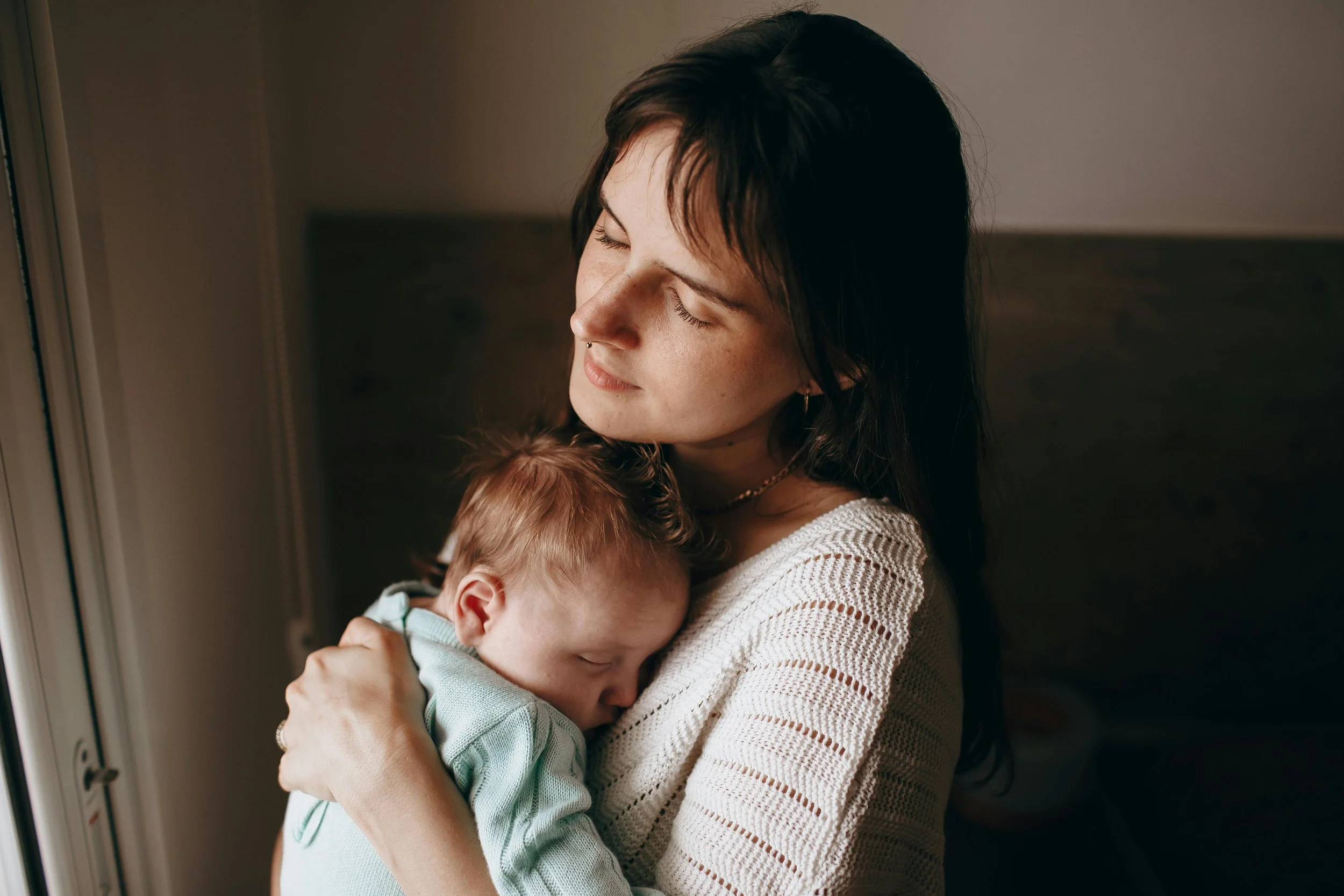 Una donna con capelli neri tiene tra le braccia un bambino con capello rosso che si appoggia al suo petto, in un momento di tenerezza.
