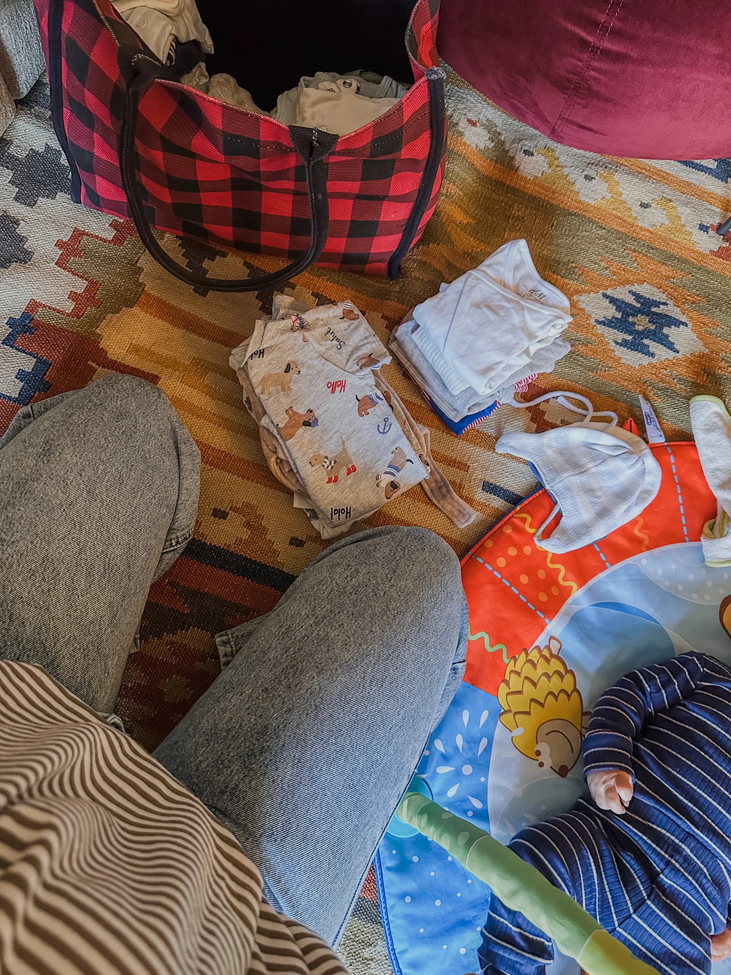 Vano con capelli curvy, piedi incrociati, tappetino colorato con una scena di un orso e un cervo, pannolini, vestiti e una borsa con un motivo a scacchi rosso e nero.