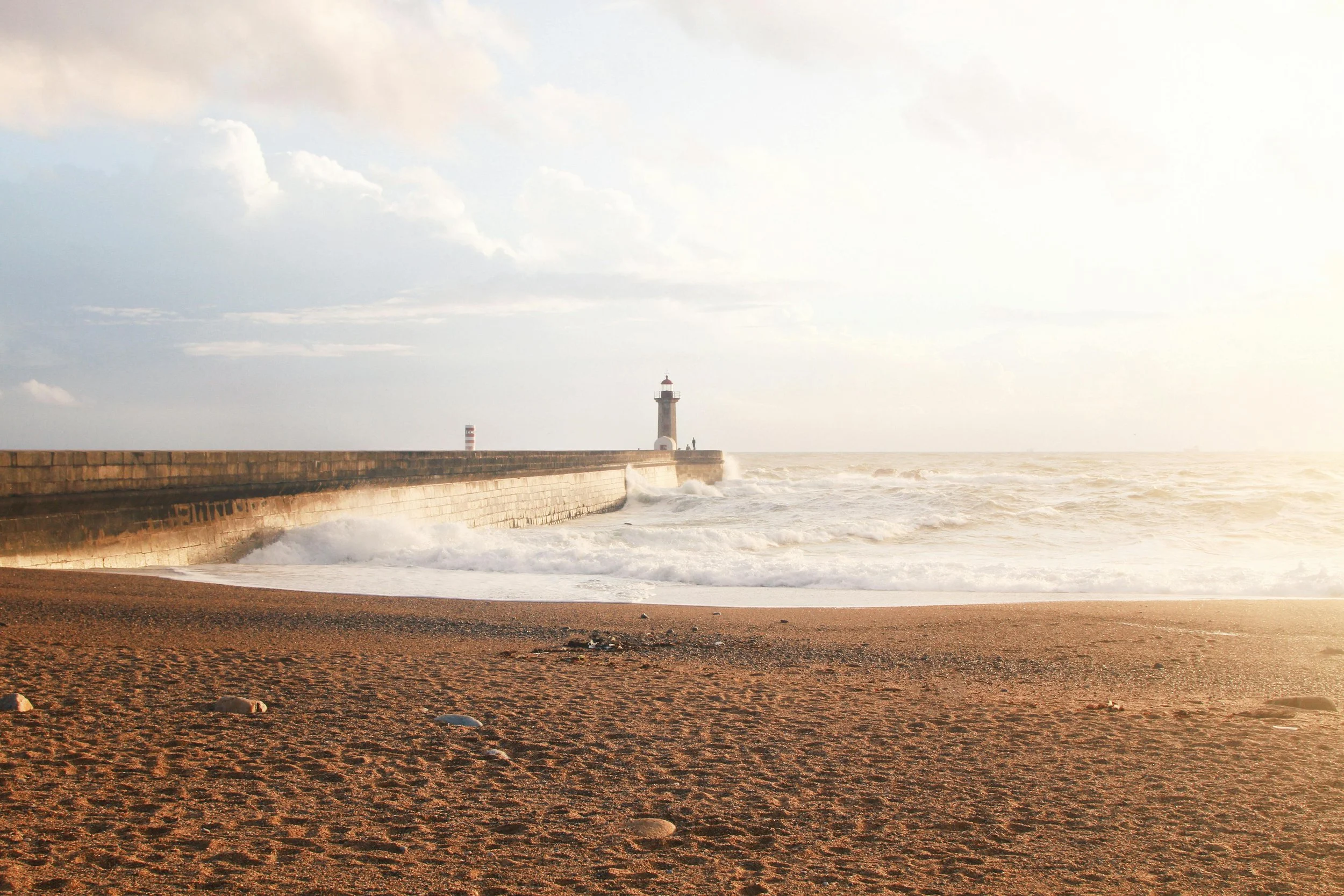 Spiaggia con sabbia e rocce, barriera in pietra e un faro sulla punta di un molo, onde che si infrangono, cielo nuvoloso con luce solare