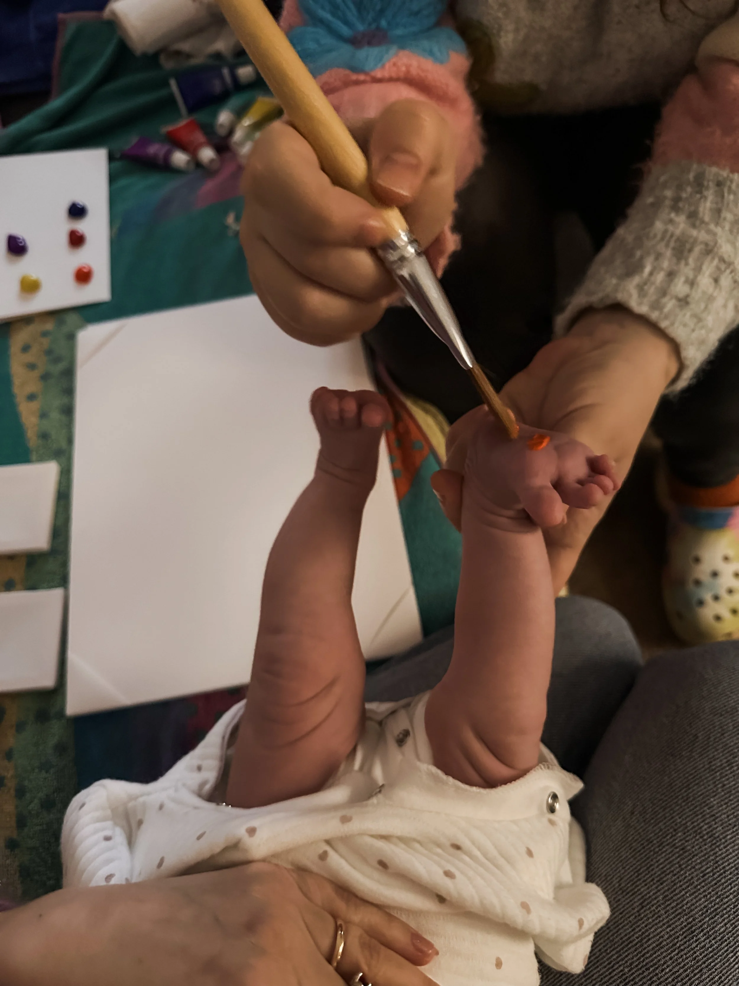Un adulto dipinge un arancione sulla mano di un neonato mentre è sdraiato su una superficie con carta bianca e strumenti artistici sul tavolo.
