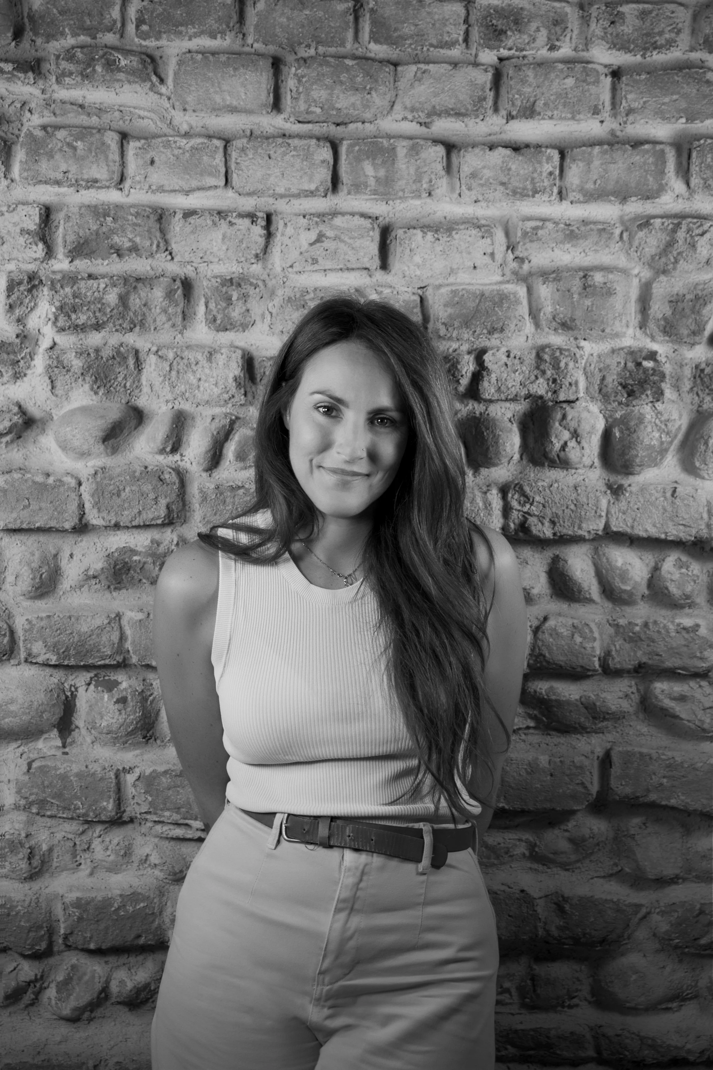 Ragazza con lunghi capelli castani, sorriso, abbigliamento casual, davanti a muro di mattoni.