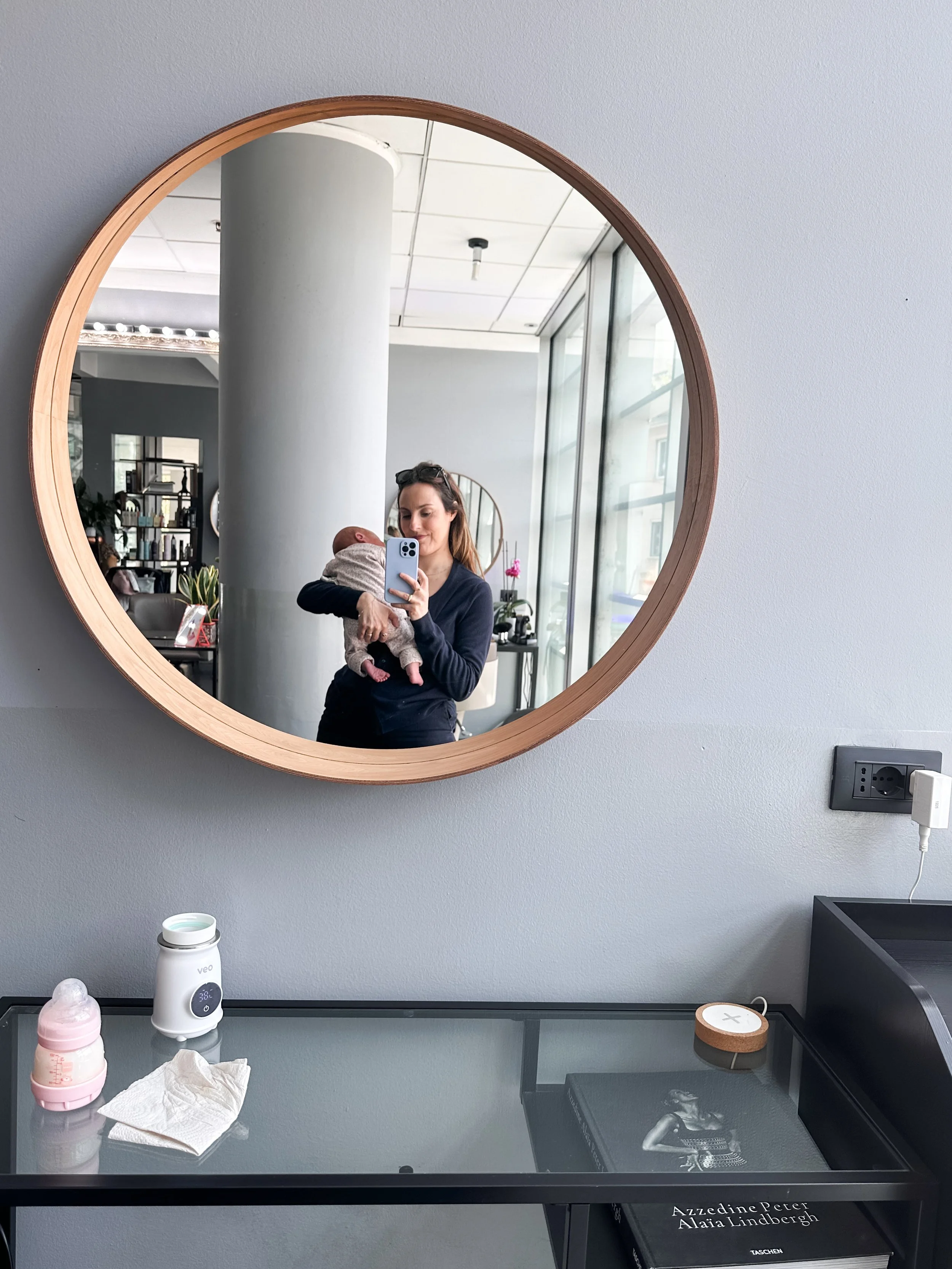 Specchio rotondo sulla parete con una donna che tiene in braccio un bambino e si sta facendo un selfie, riflesso dell'interno di un locale con colonne e scaffali.