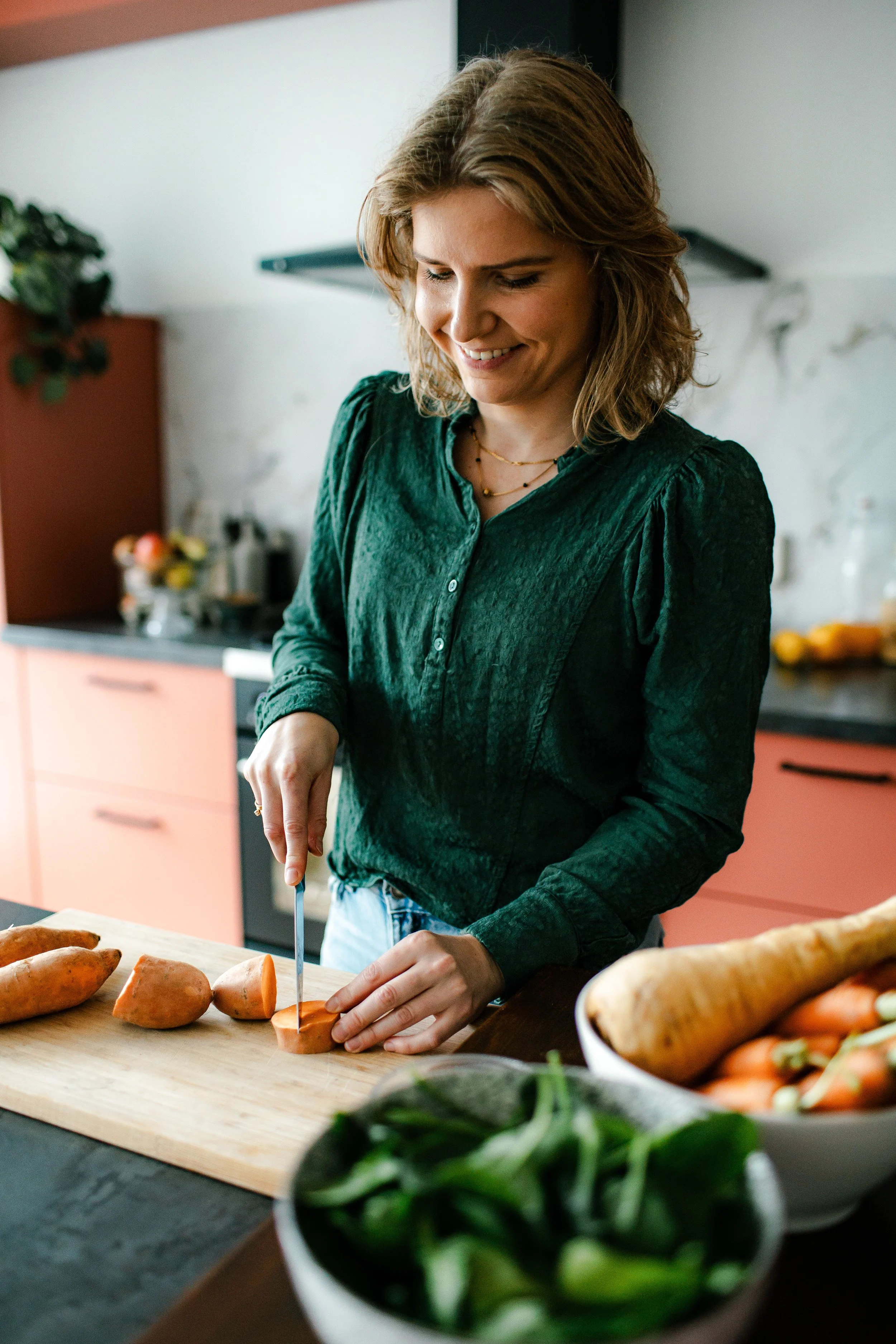 A woman in a green blouse slices a sweet potato on a wooden cutting board in a modern kitchen.