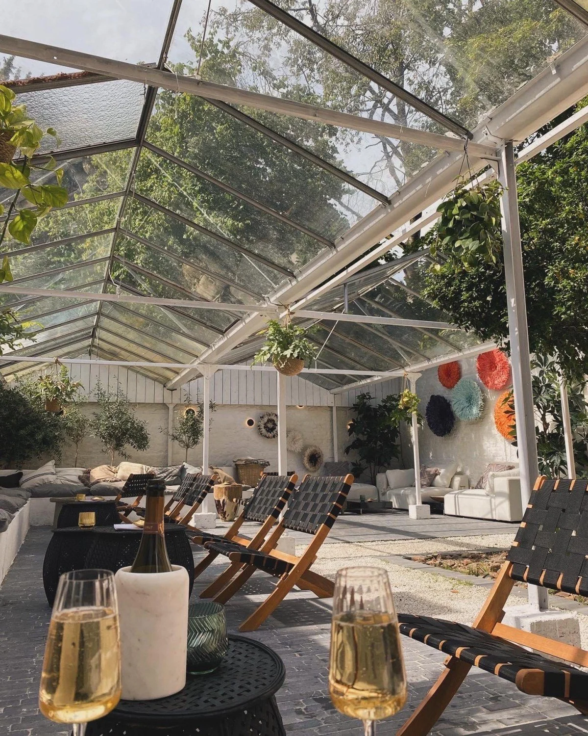 Gedeelte van een gezellige buiten lounge met houten stoelen, een salontafel met drankjes, een glazen kas met planten en kleurrijke decoraties aan de muur.