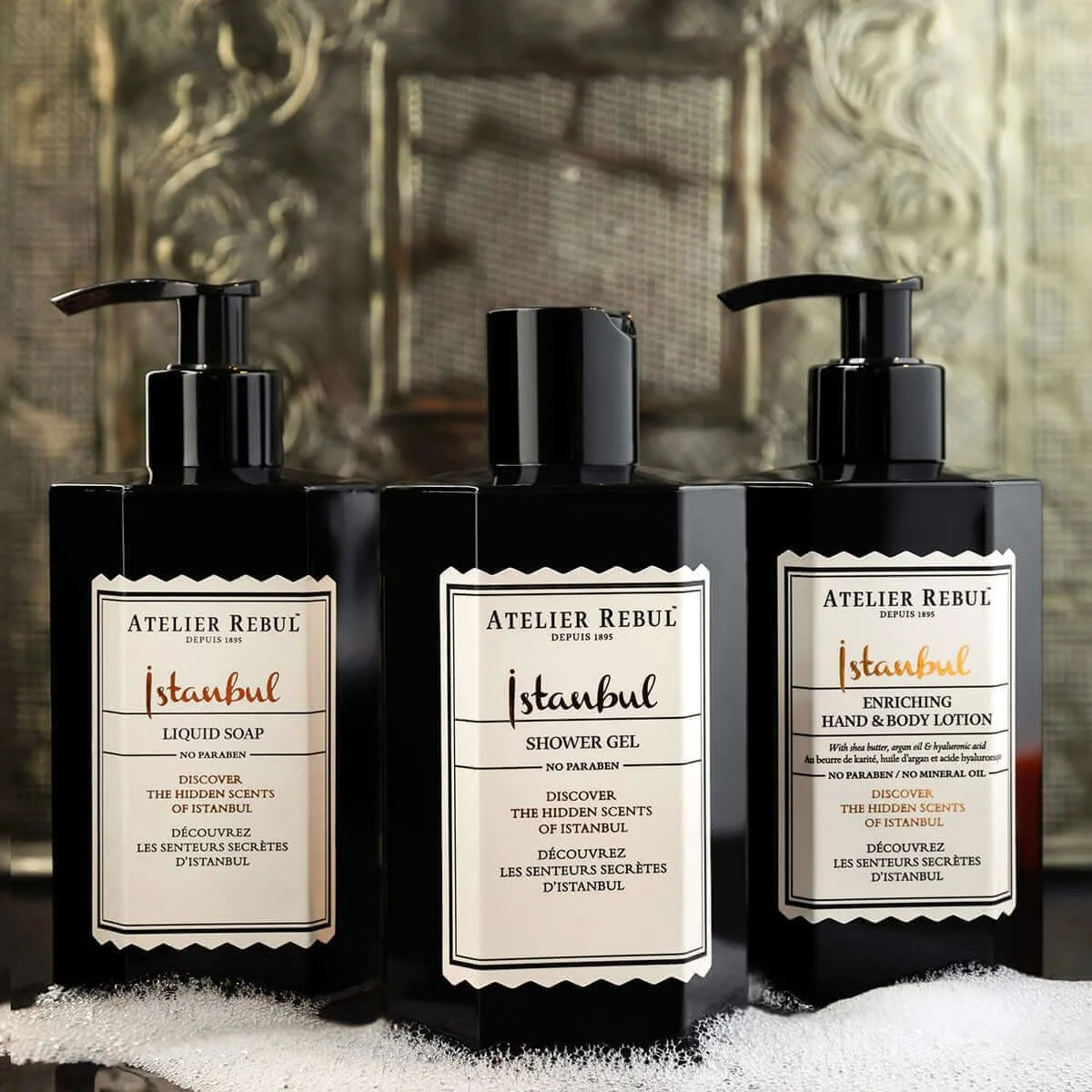 Drie zwarte flessen met verzorgingsproducten van het merk Atelier Rebul voor Istanbul, waaronder liquid soap, shower gel en hand- en bodylotion, geplaatst op een wit oppervlak met een decoratieve achtergrond.