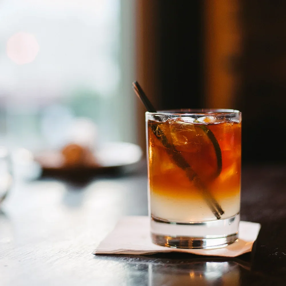 Een glas met een koud drankje, waarschijnlijk een cocktail, met ijsblokjes, een citroen- of limoenpartje en een rietje, geplaatst op een witte onderzetter op een houten tafel.