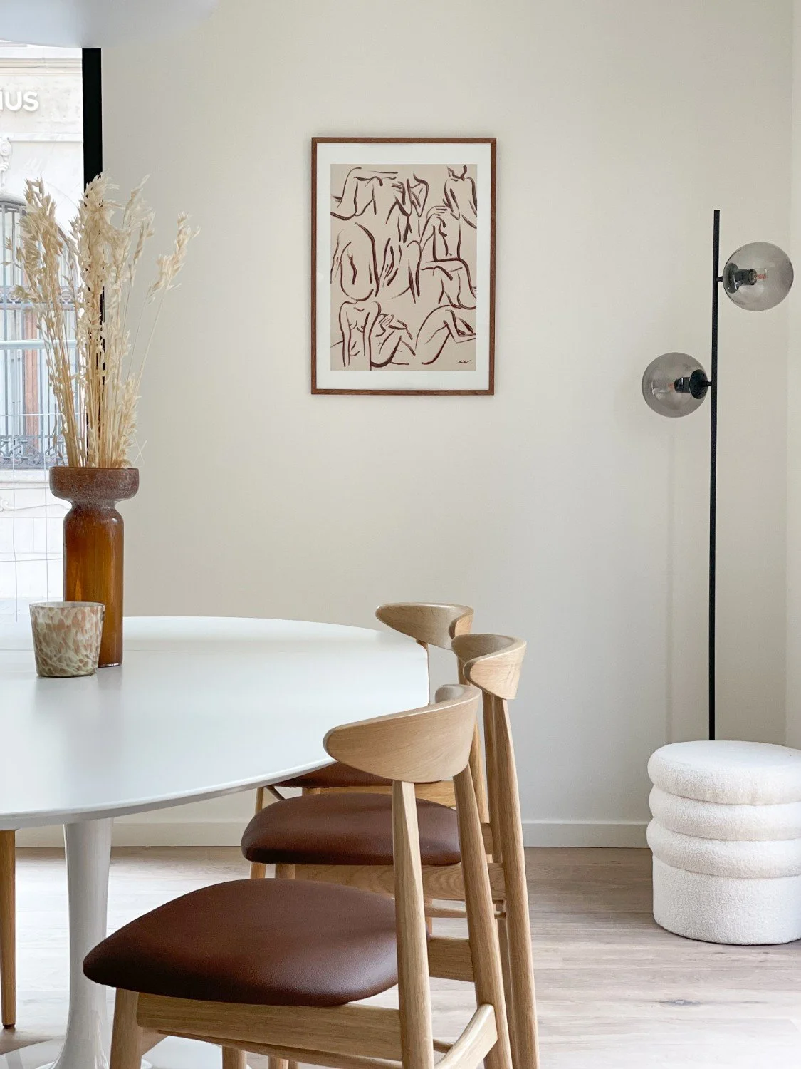 Interieur van een modern eetkamer met een witte ronde tafel, houten stoelen met leren zittaken, een vaas met gedroogde bloemen, een muurschildering van abstracte figuren, een vloerlamp met twee glazen bollen en een gestapelde witte poef.