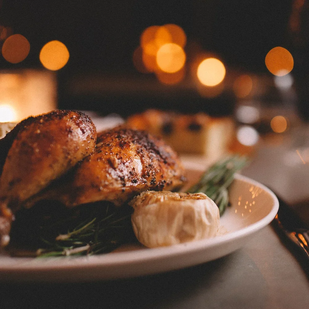 Geroosterde kip met knoflook en groenten op een witte schaal, met achtergrond van vuurbouwen en lichten in de duisternis.