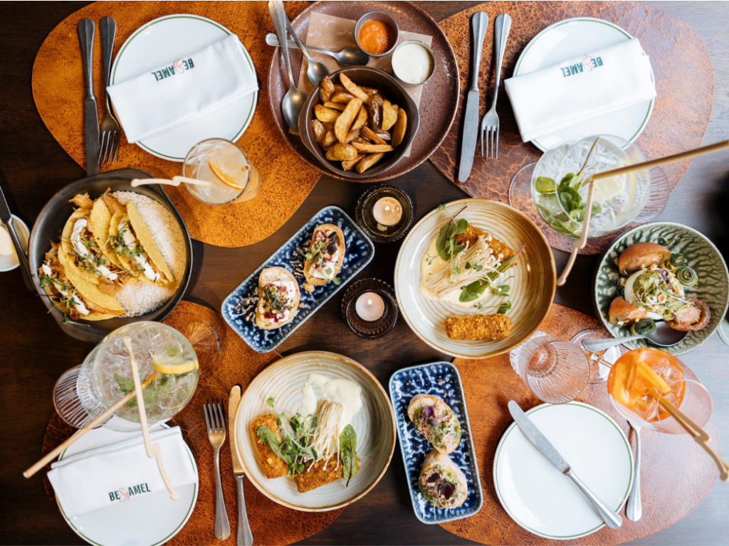 Een tafel met diverse gerechten, waaronder taco's, sandwiches, salades, en drankjes, op houten onderzetters.